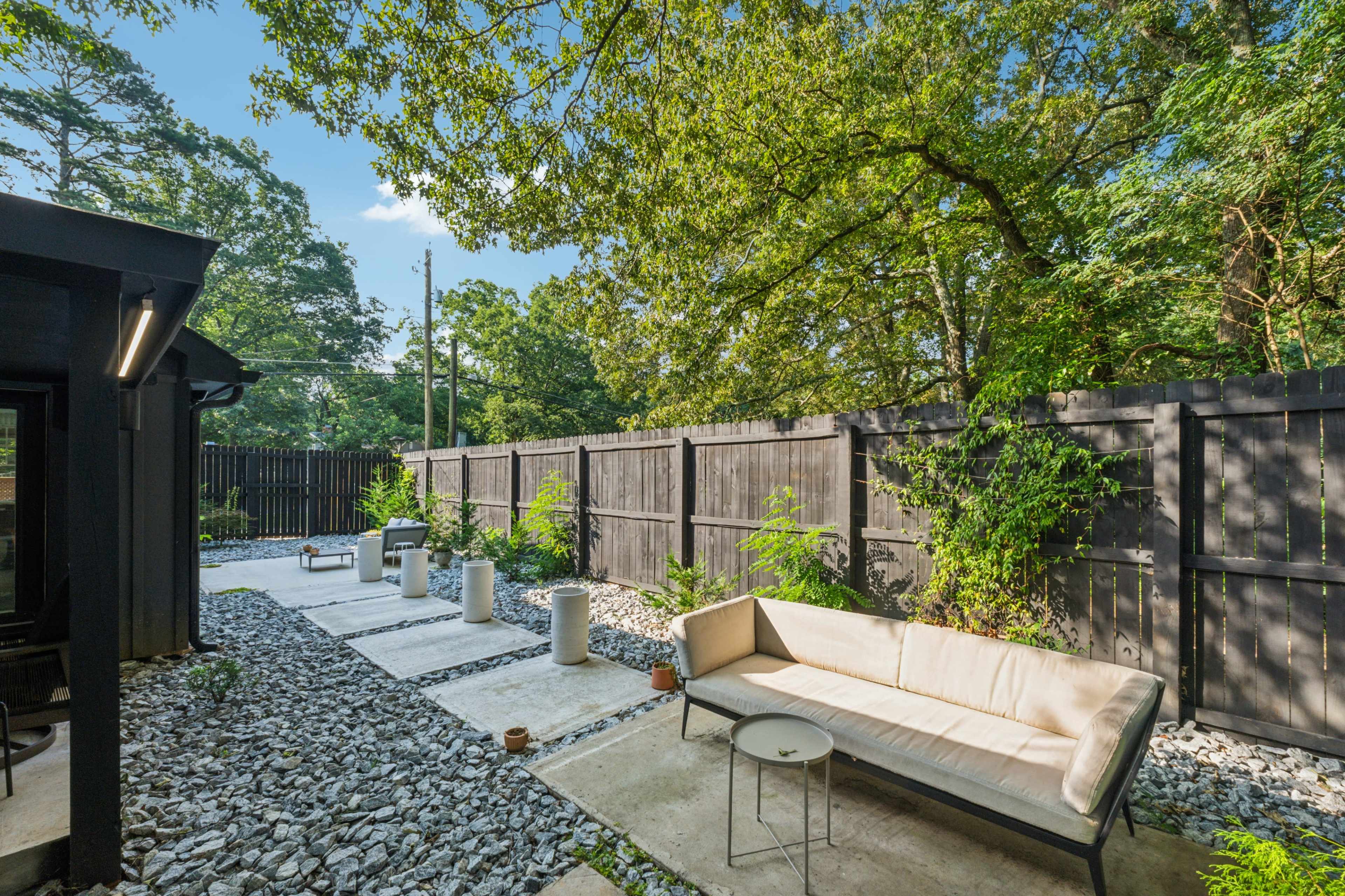 The image shows a patio area with a gravel path, concrete slabs, a sofa, and potted plants, surrounded by a wooden fence and trees.