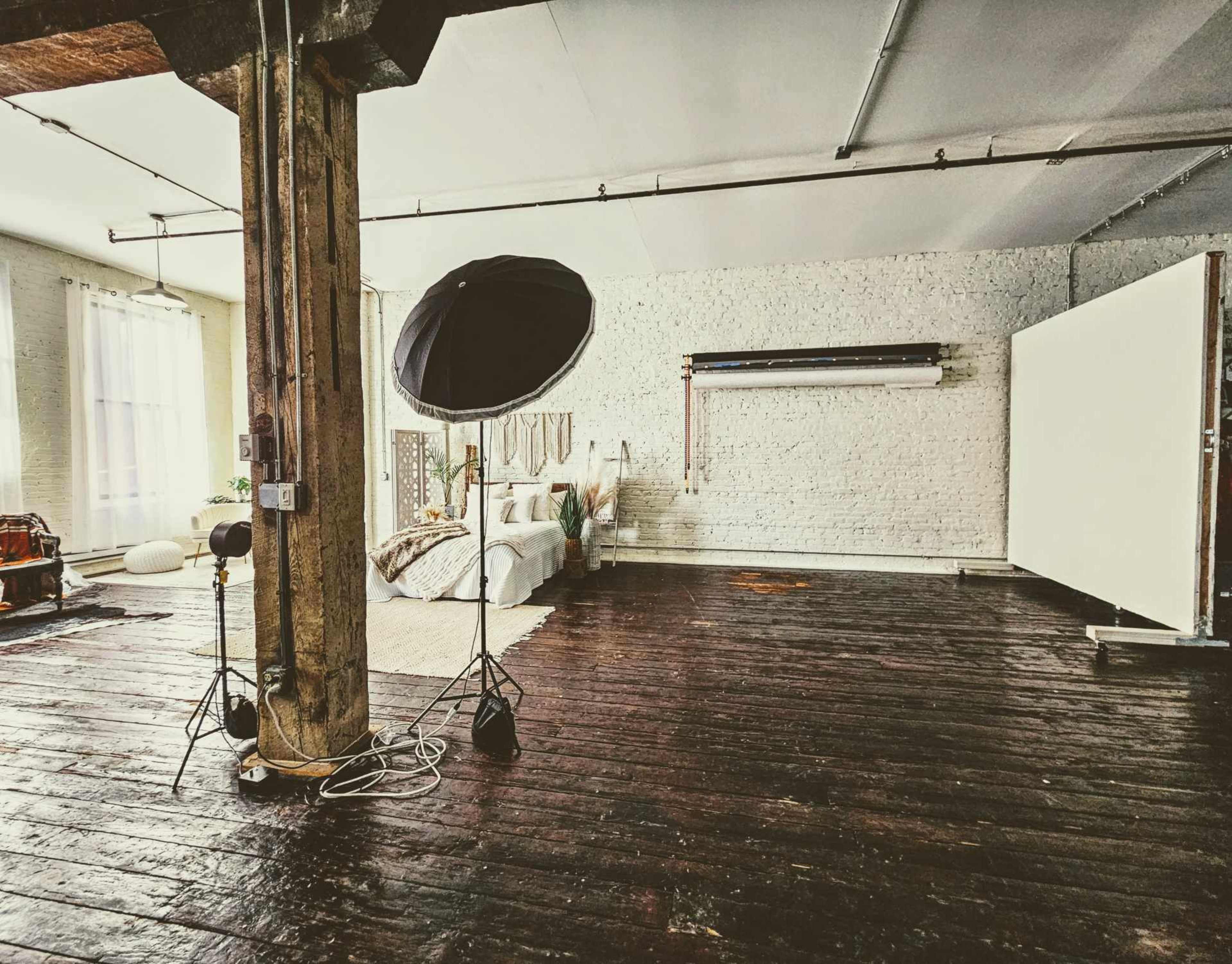 The image shows a spacious, well-lit bedroom with wooden floors, a large bed, and a photography setup including a softbox and a backdrop.