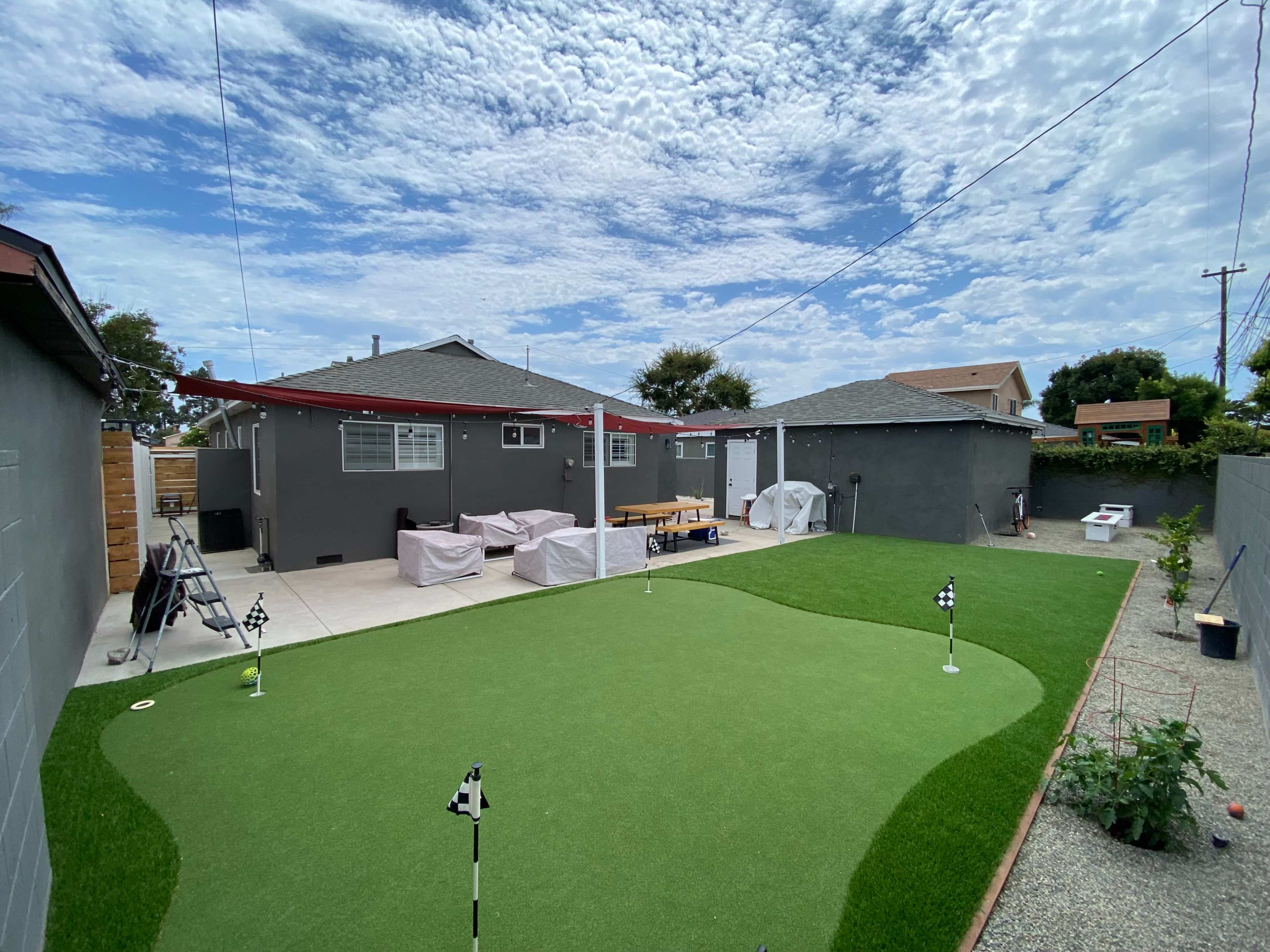 A backyard setting with a synthetic putting green, a patio area, and a gray house in the background under a partly cloudy sky.