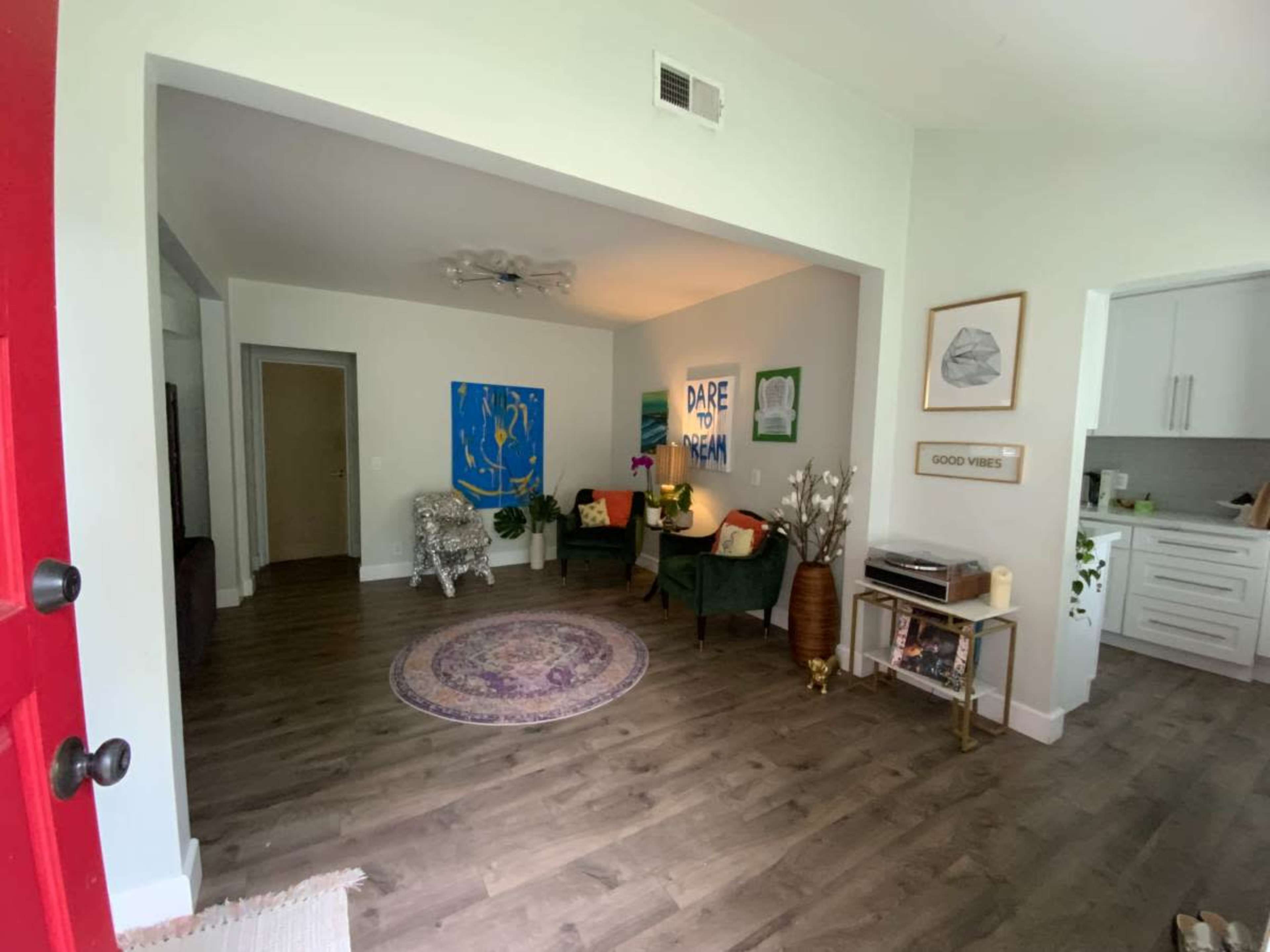 The image shows a bright, modern living room with a circular rug, two green chairs, and artwork on the walls.