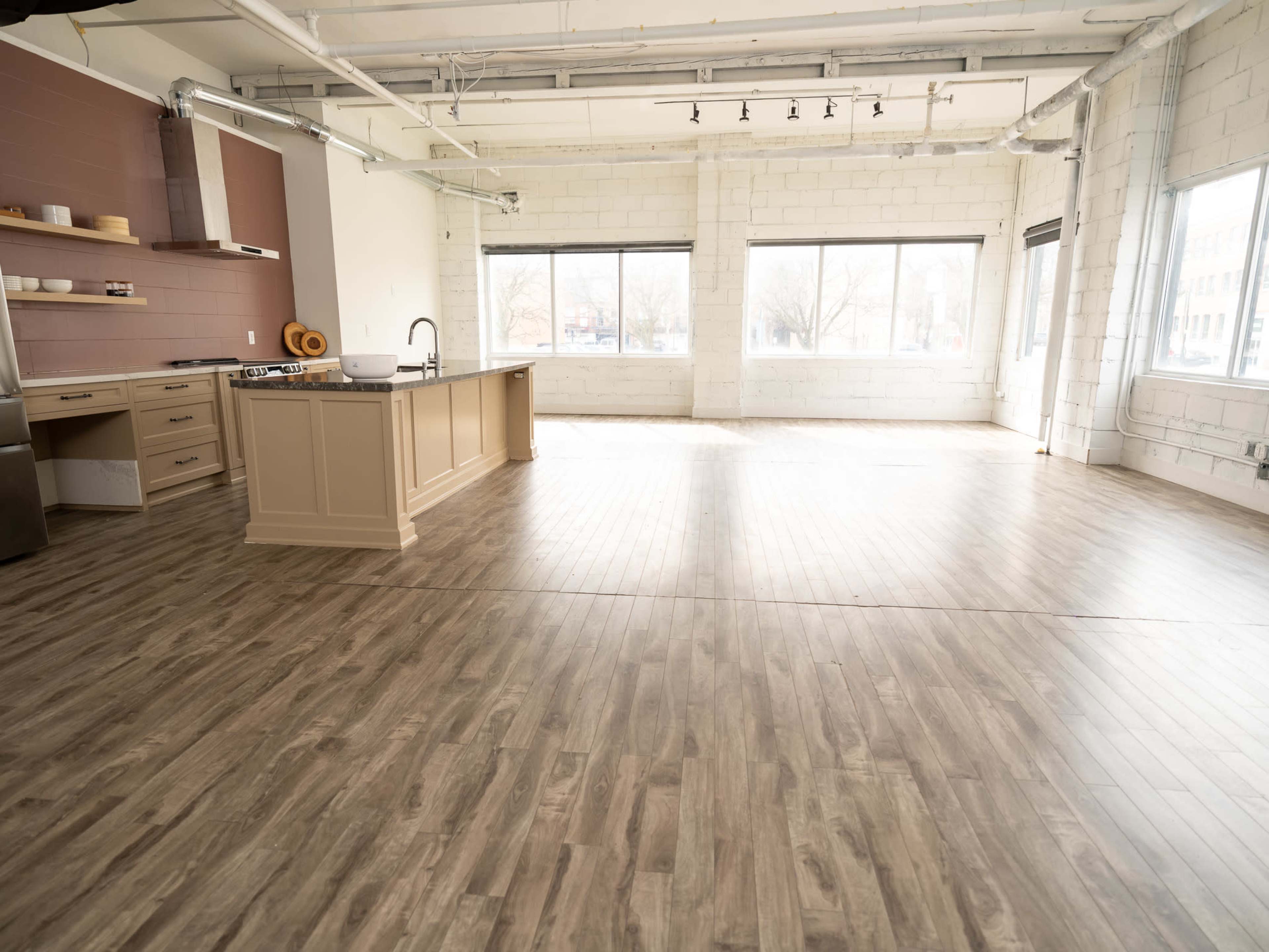 The image shows a spacious, modern kitchen area with a large island, featuring windows that allow natural light to fill the room and a wooden floor.