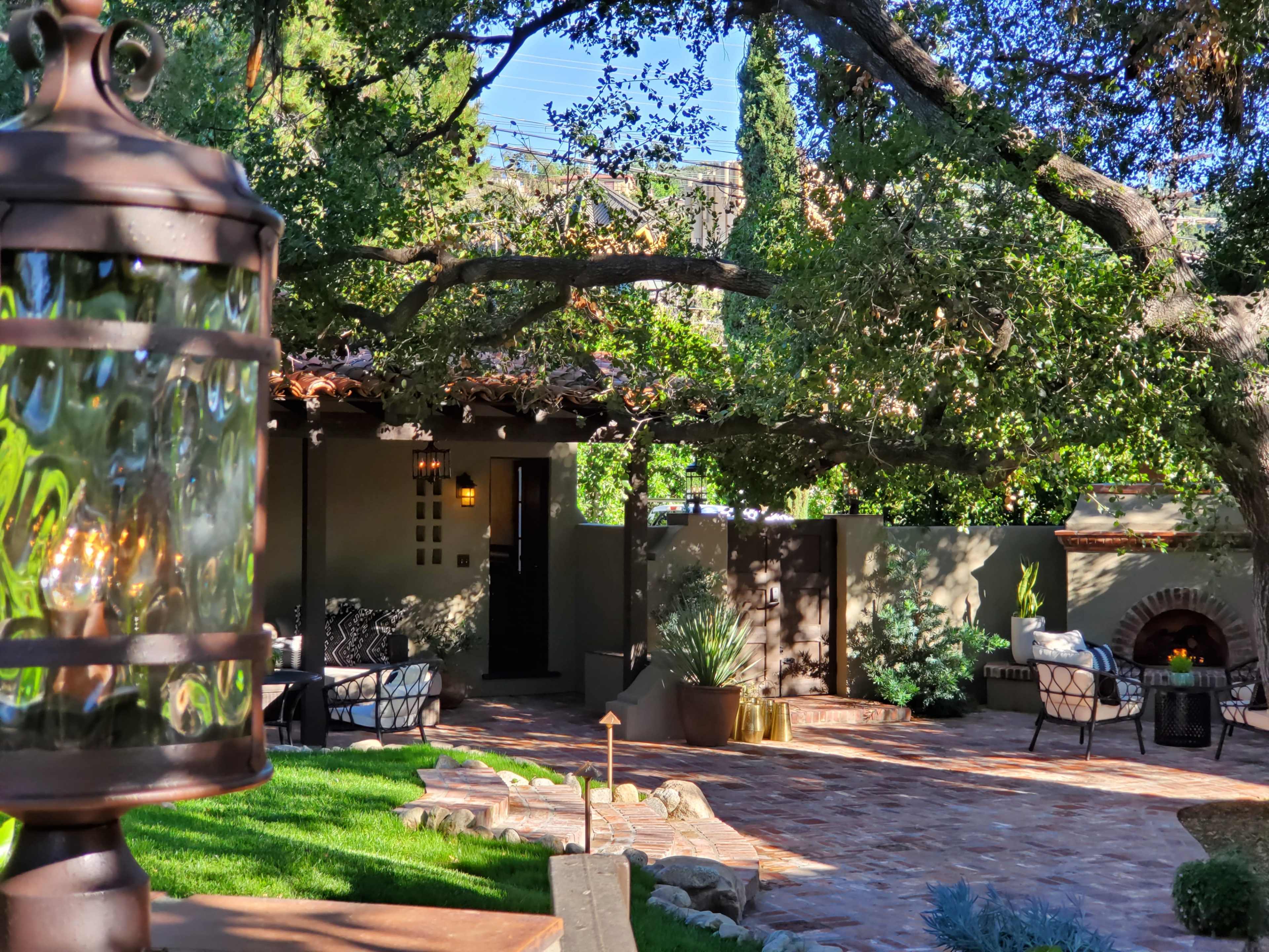 A well-landscaped outdoor patio features lush greenery, a stone path, and seating arrangements under a canopy of trees.