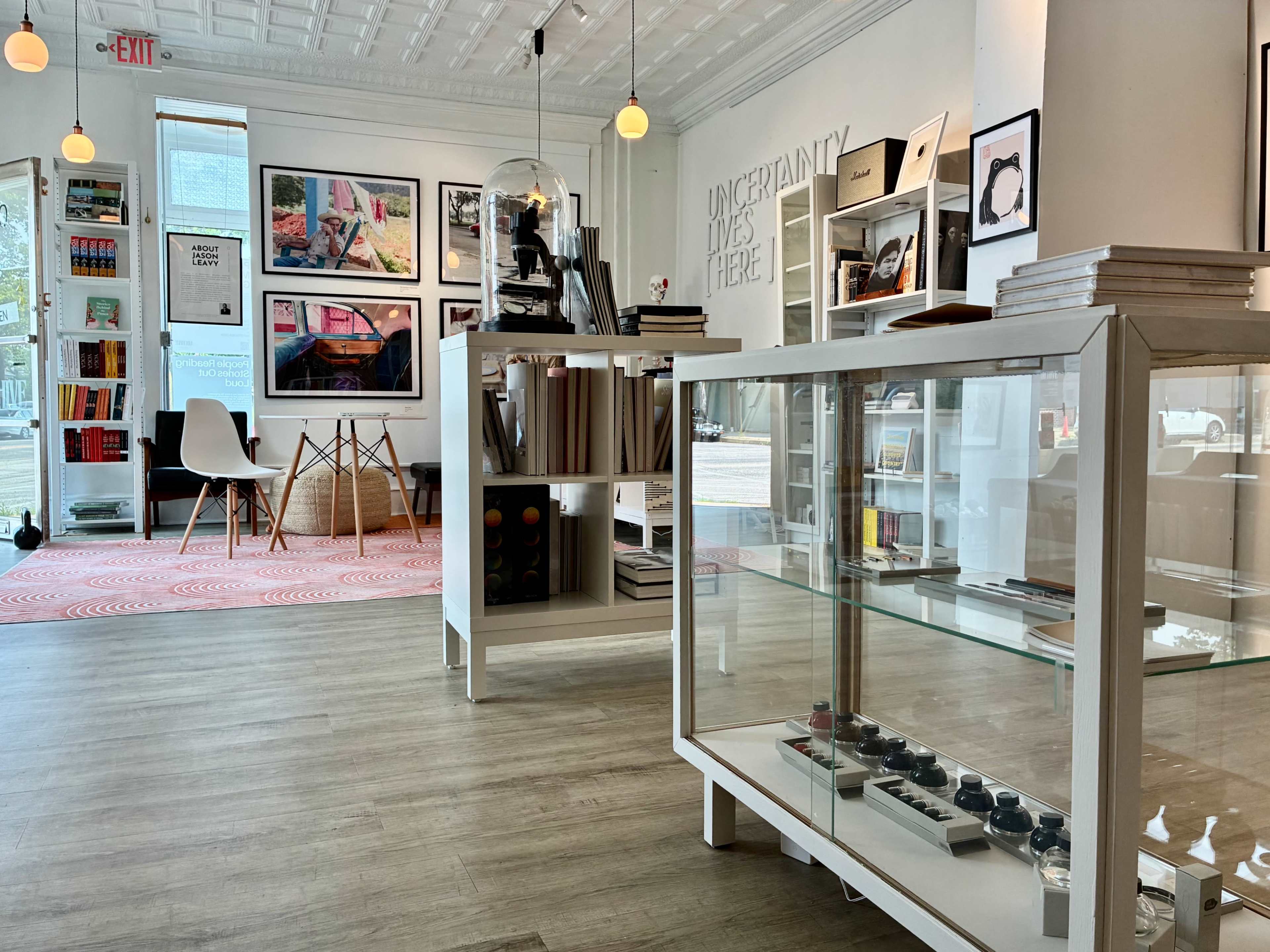 The image shows a modern, well-lit interior of a creative space featuring bookshelves, artwork on the walls, and display cases with various items for sale.