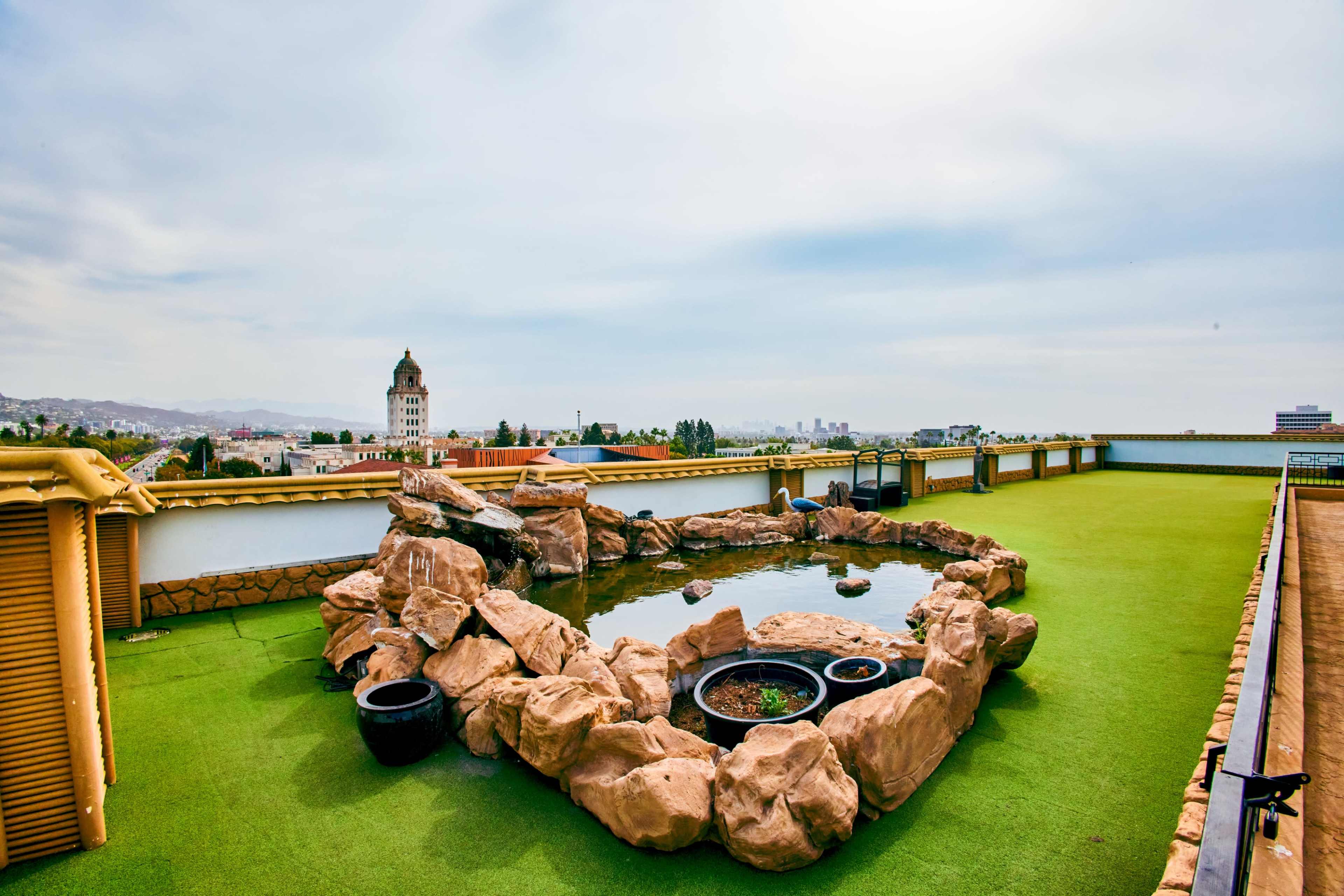 The image shows a rooftop terrace featuring a rock formation surrounding a pond, with a view of a cityscape in the background.