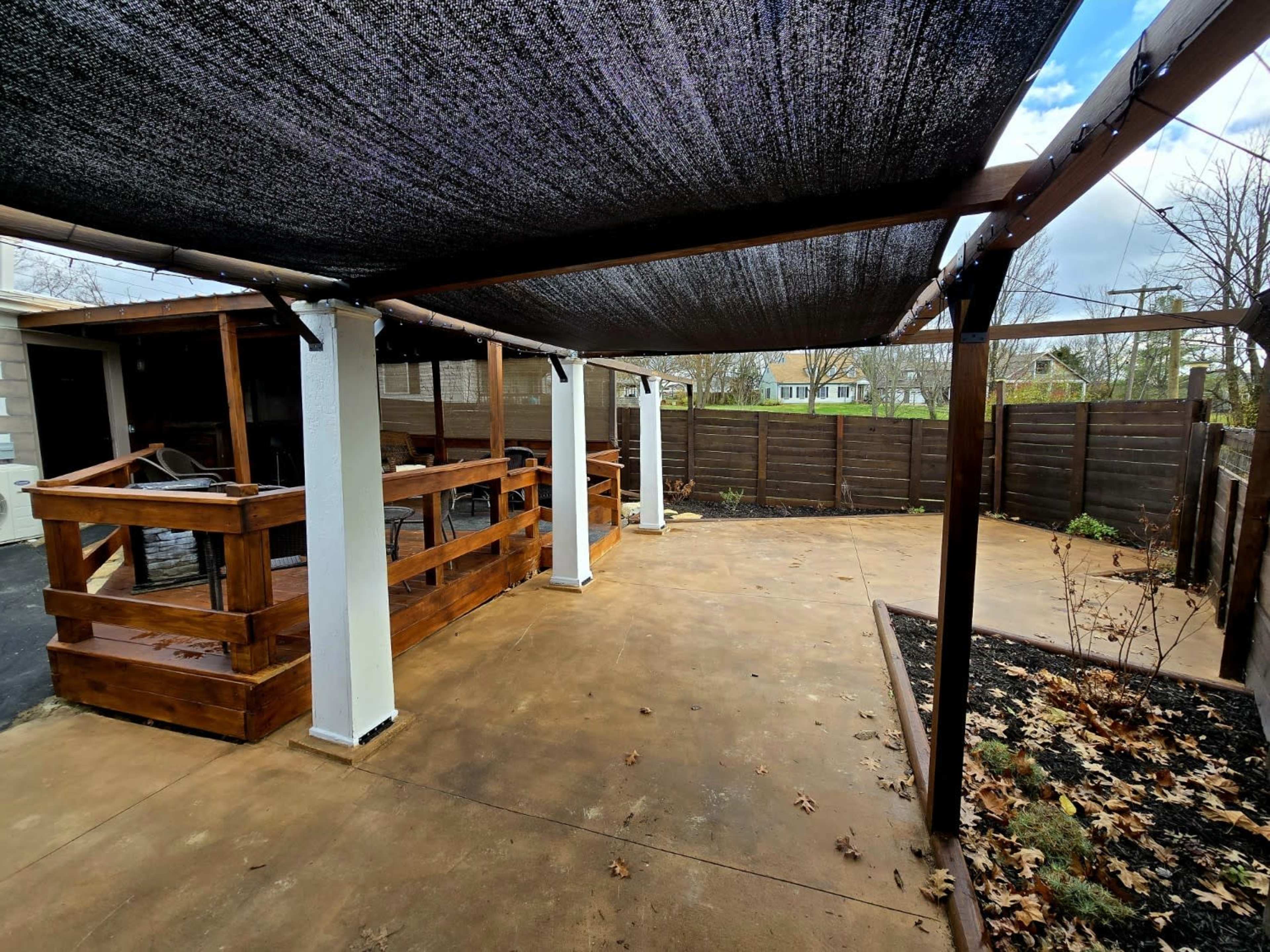 The image shows a covered outdoor patio area with a concrete floor, wooden seating structures, and a section of a fenced garden.