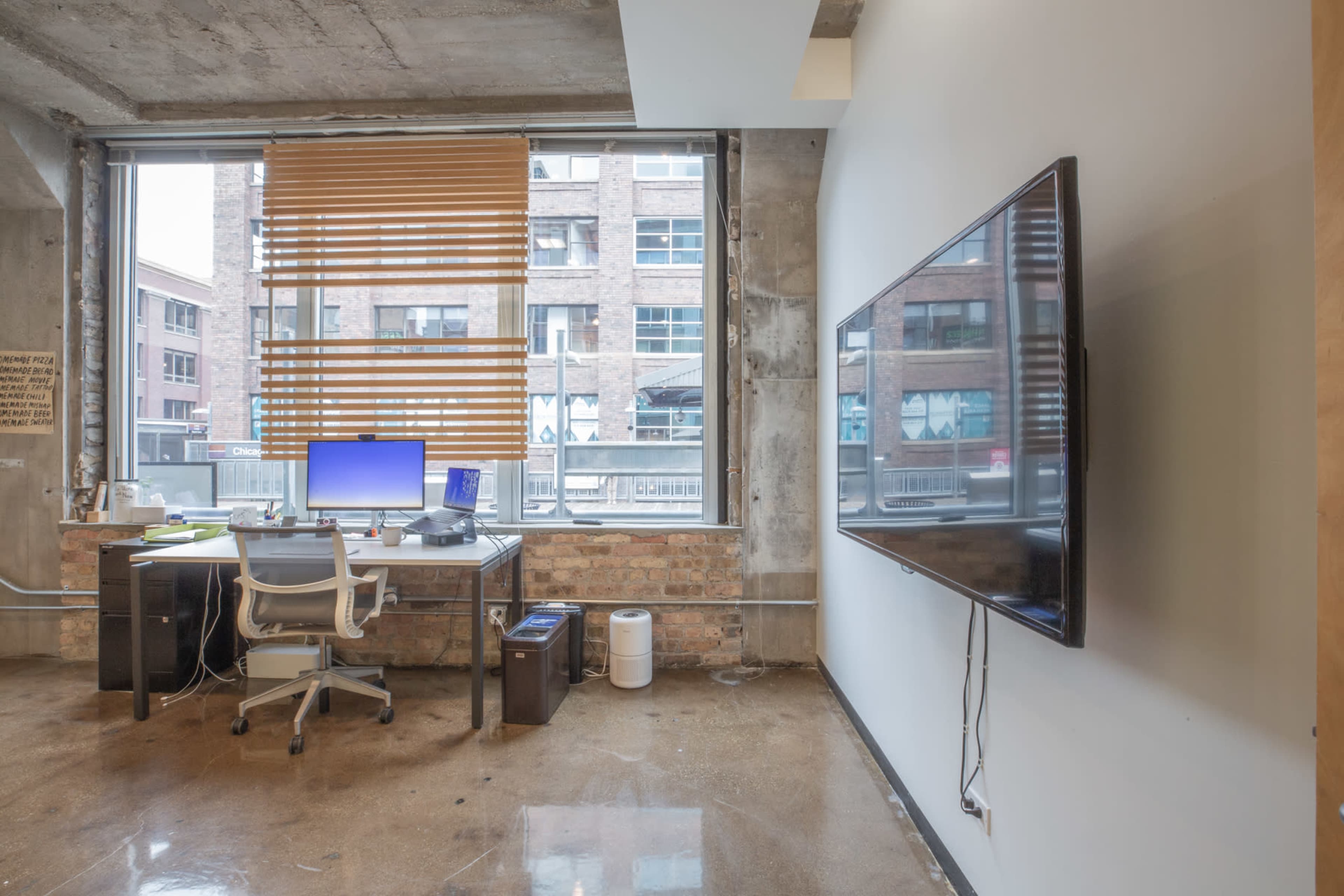 The image shows an office space featuring a desk with a computer, a television mounted on the wall, and large windows with wooden blinds, revealing an urban setting outside.