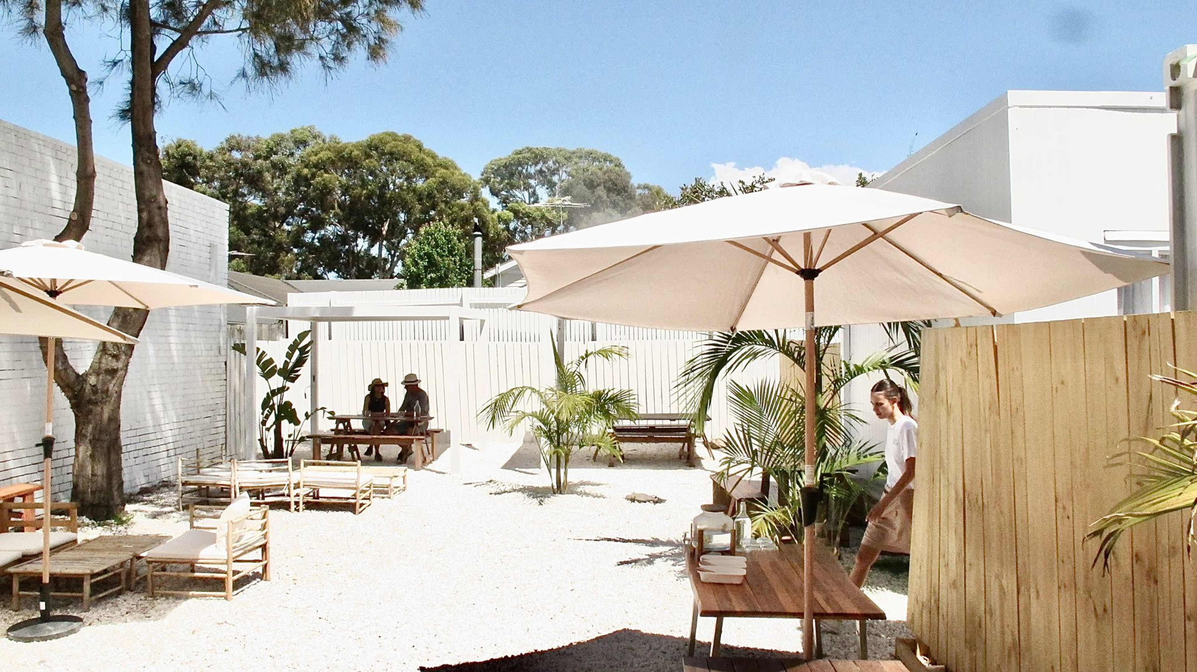 An outdoor seating area with wooden tables, umbrellas, and lounge chairs arranged among gravel and greenery.