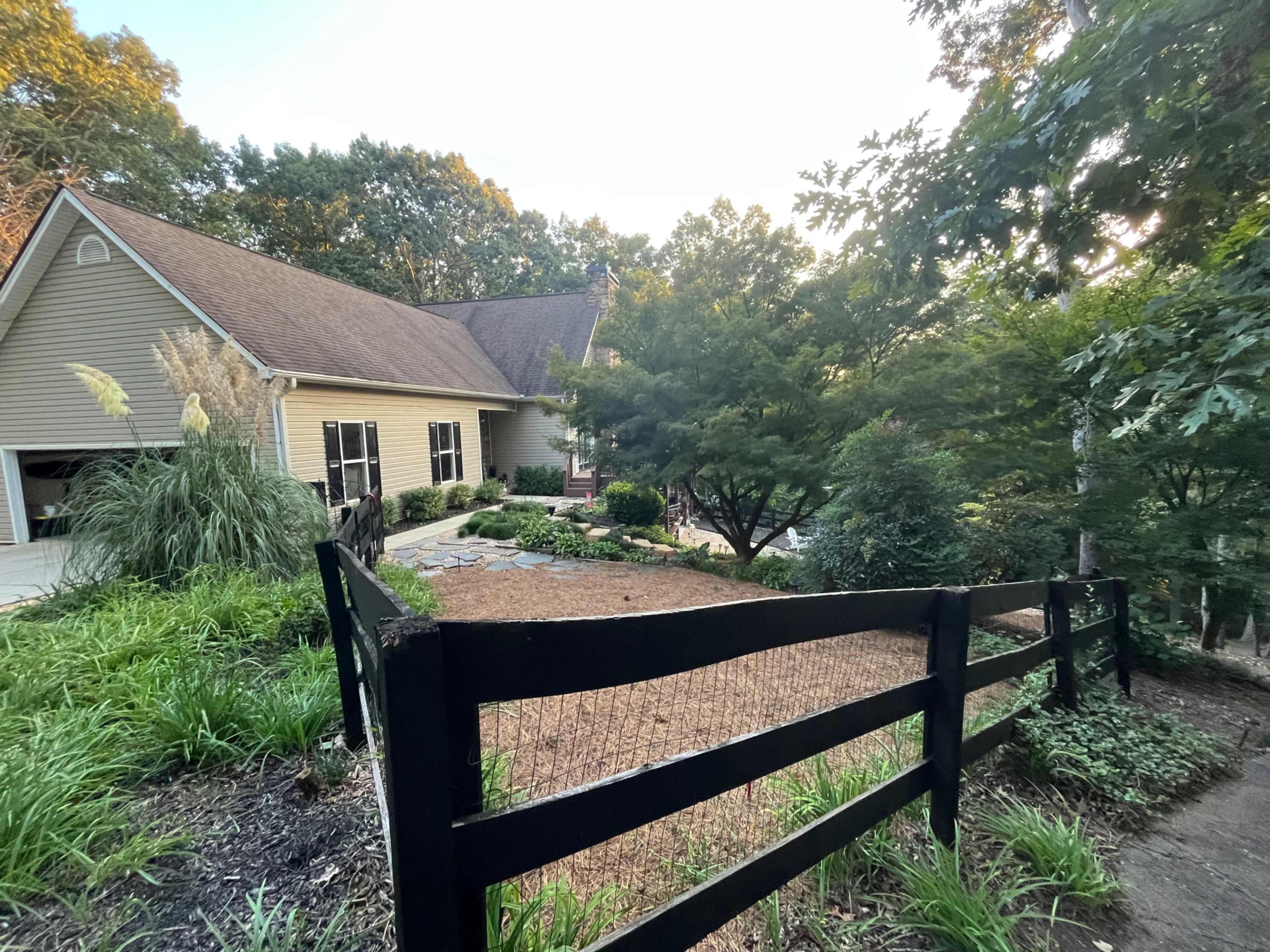 A house with a sloped yard is surrounded by trees and a black wooden fence, leading to a gravel pathway and garden.