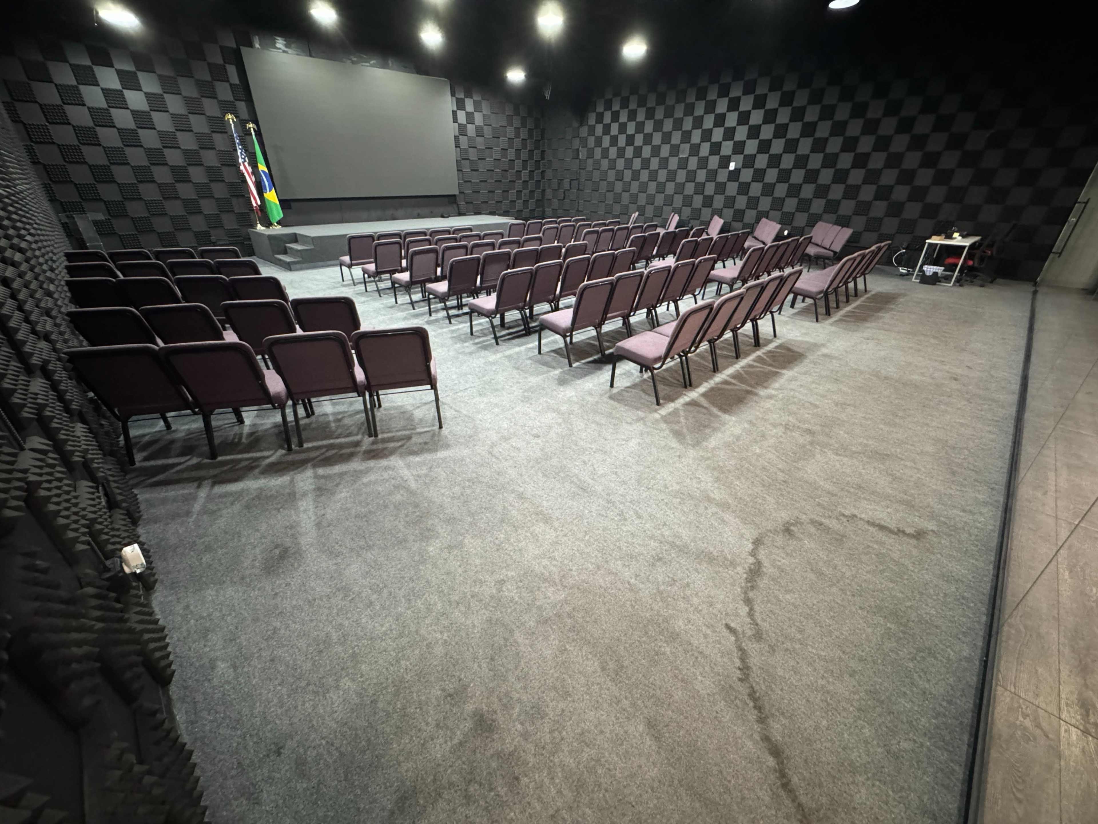The image shows a nearly empty auditorium with rows of purple chairs arranged facing a large screen, flanked by a Brazilian flag in the background.