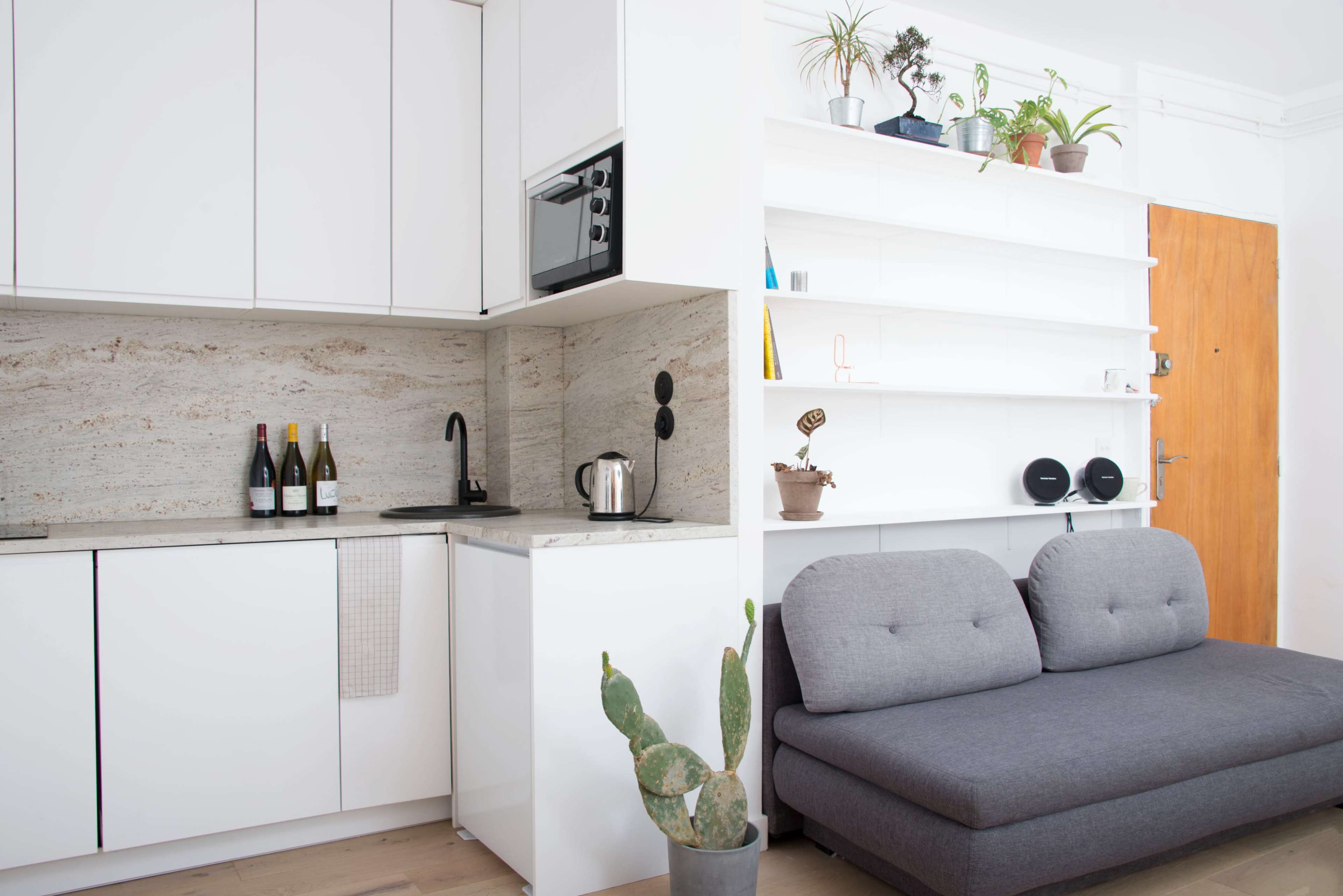 The image shows a modern kitchen with white cabinetry, a marble backsplash, and a gray sofa facing a white shelf with plants and speakers.