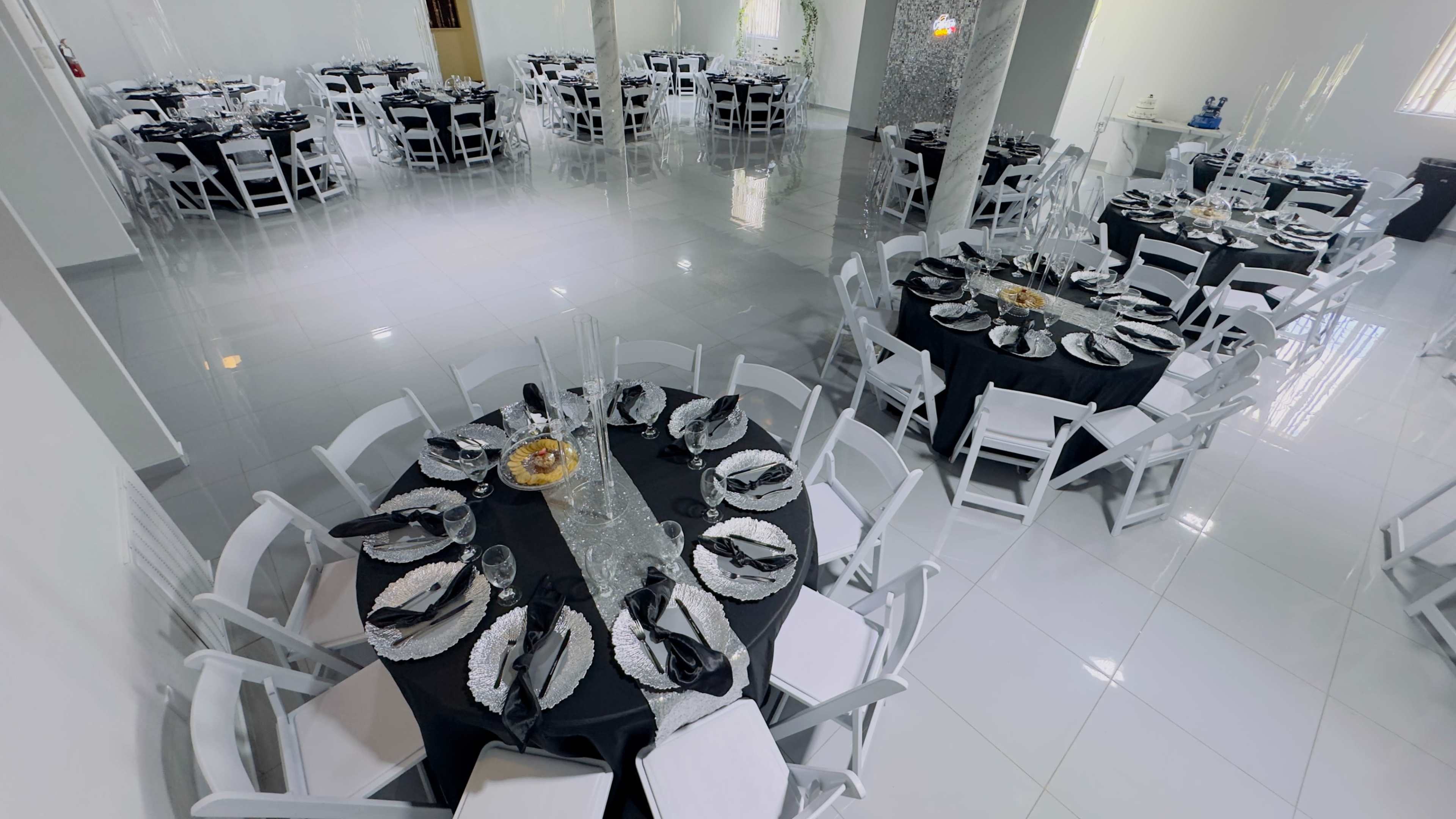 The image shows a large banquet hall set up with multiple round tables covered in black tablecloths, adorned with silver tableware and centerpiece decorations.