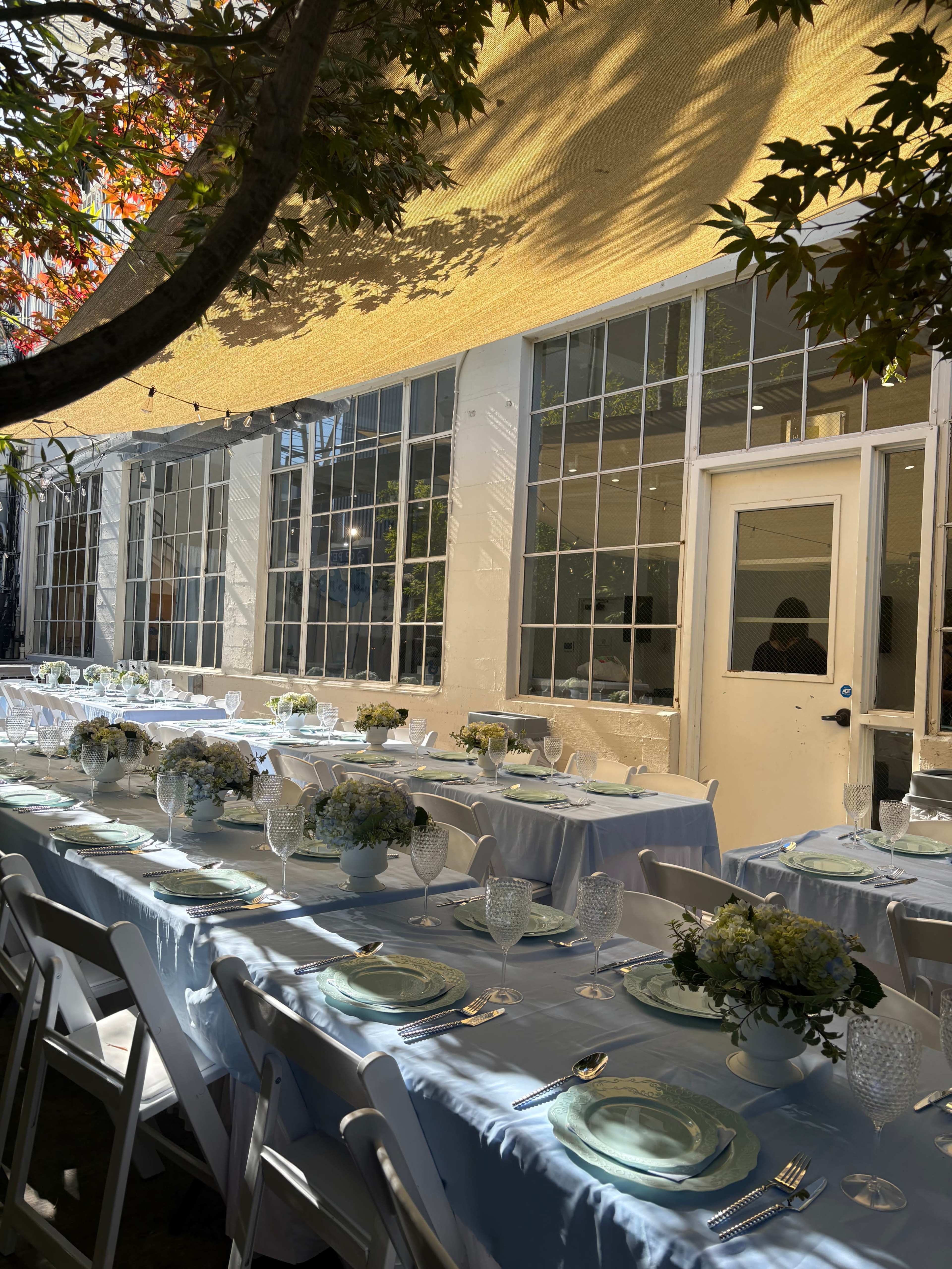 A long dining table is set with glasses, plates, and floral centerpieces under a canopy of trees, next to large windows that let in natural light.