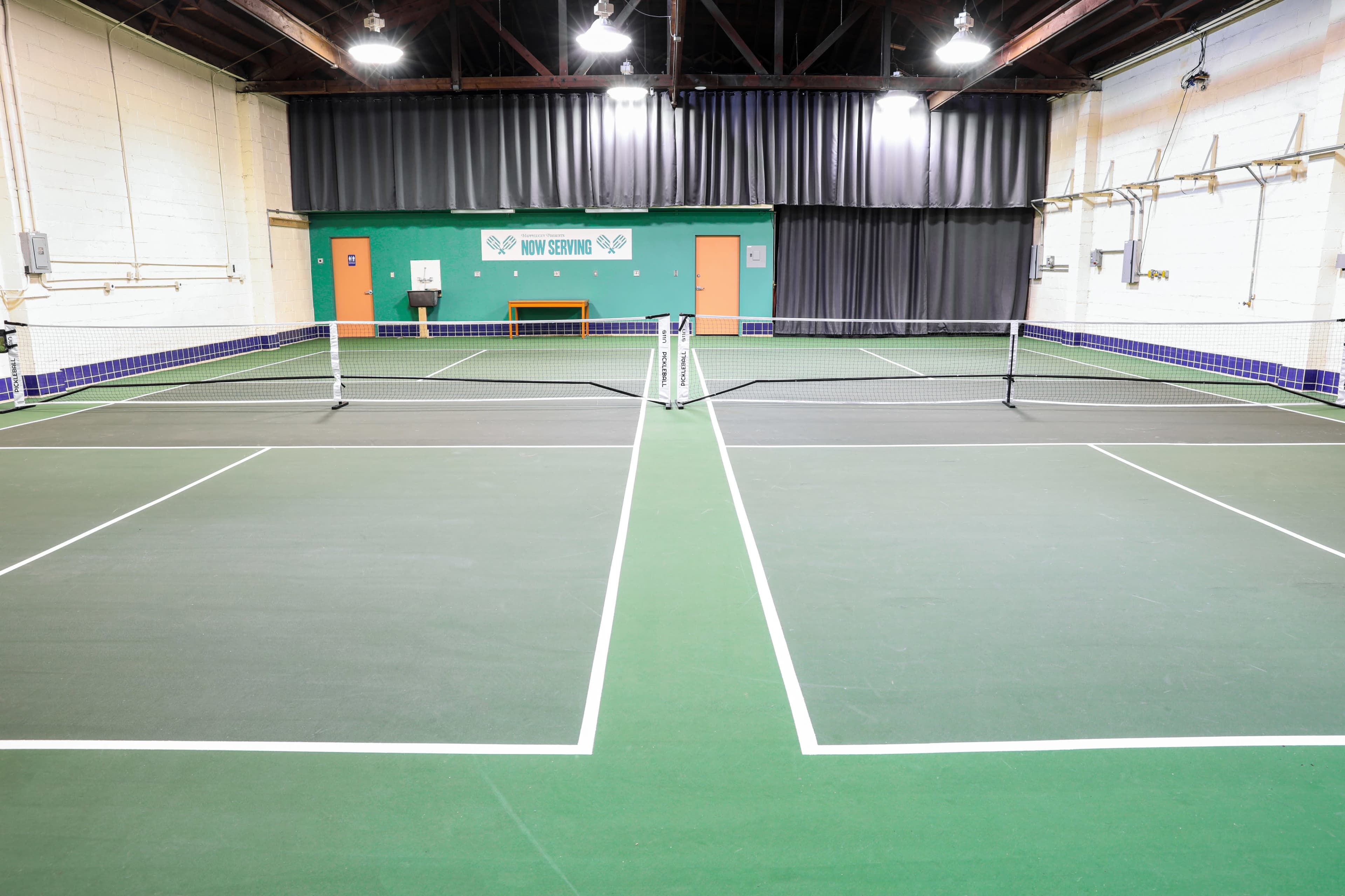 The image depicts an indoor tennis court with a green playing surface, complete with netting and surrounding walls.