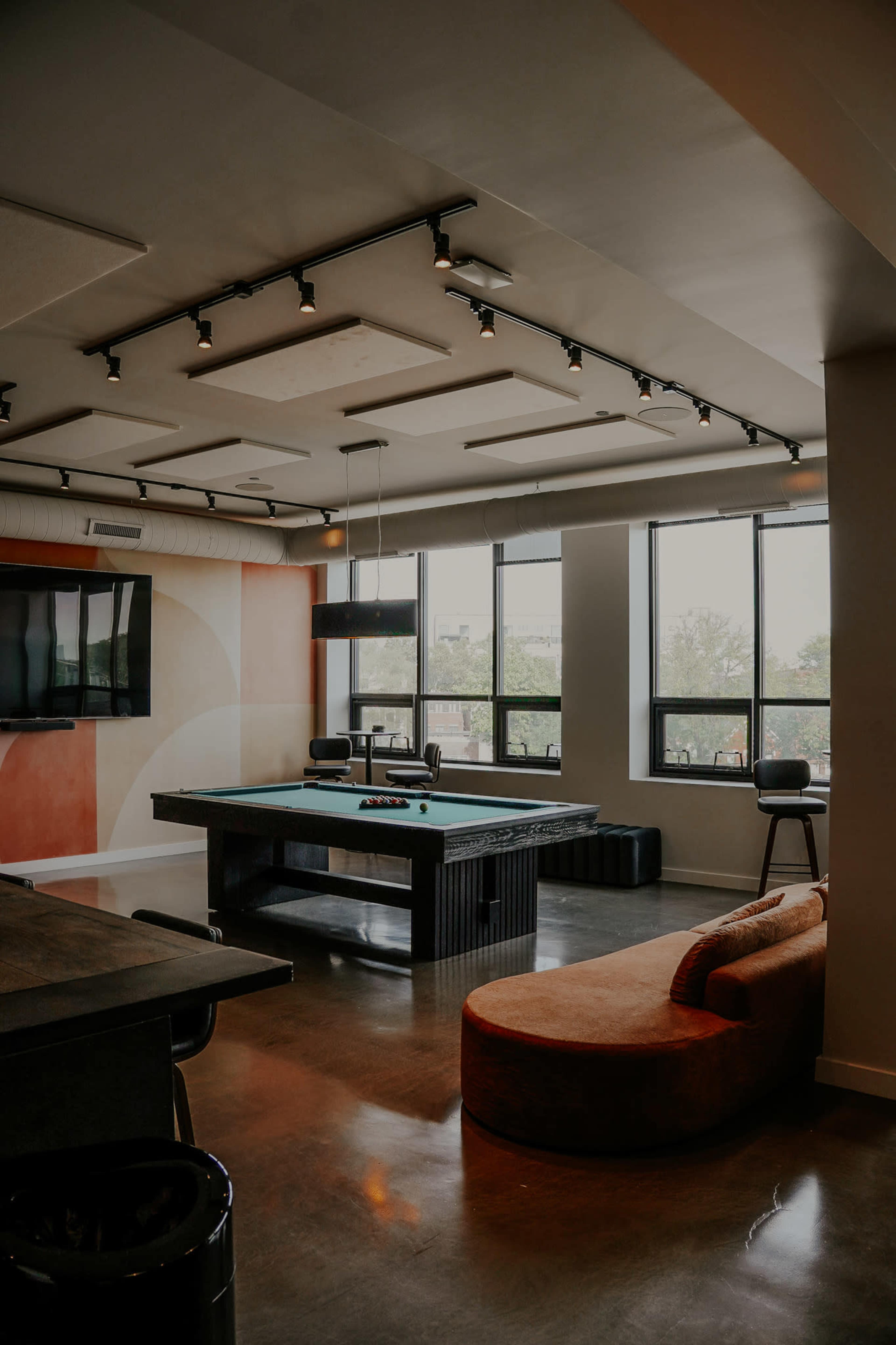 The image shows a modern game room featuring a pool table, lounge seating, and large windows that provide natural light.