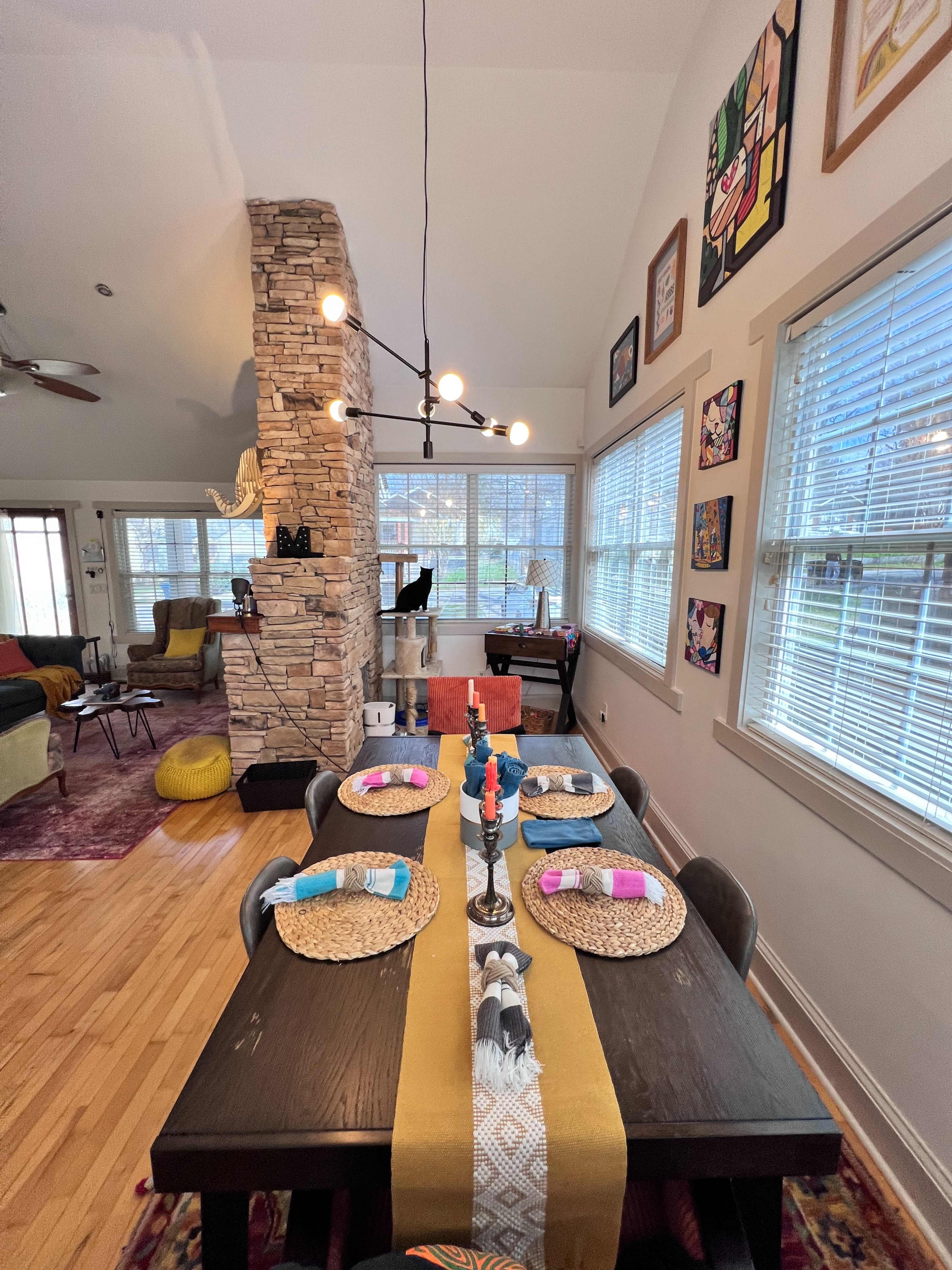 A dining table set with place settings and decorative items is positioned beside a stone column in a brightly lit room featuring large windows and various artworks on the walls.