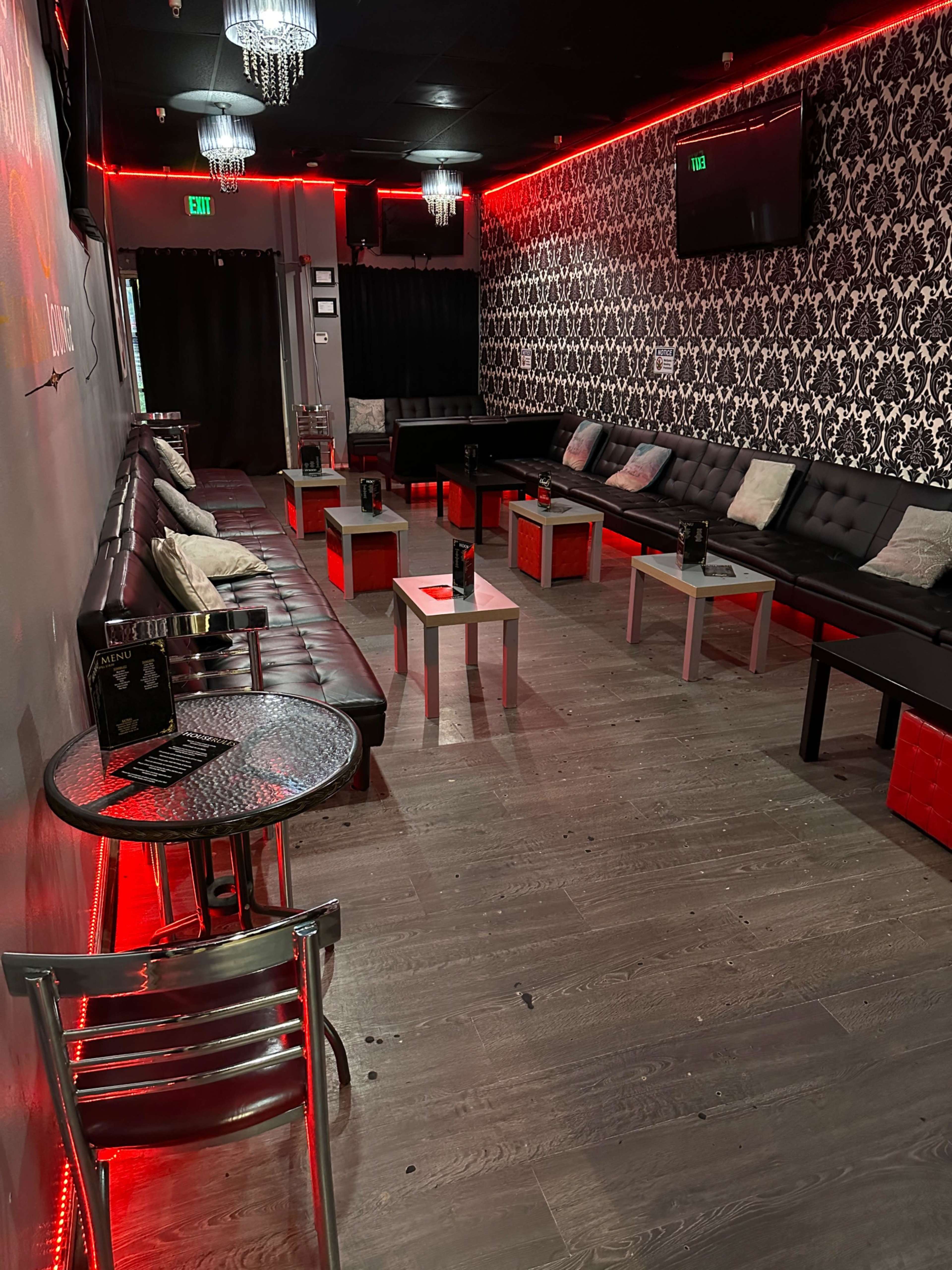 The image shows a dimly lit lounge area with black leather seating, small tables, and red neon lighting along the walls.