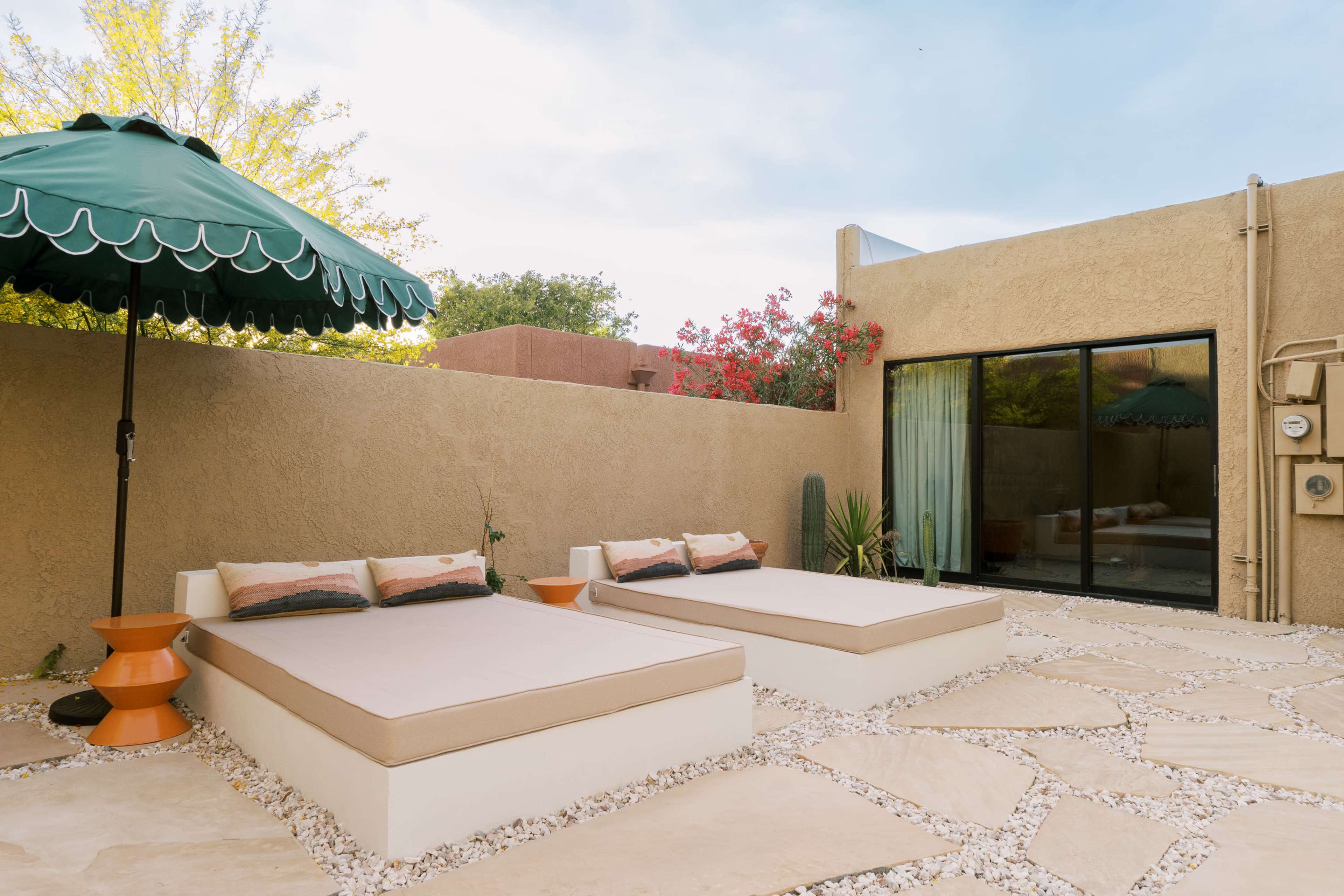 A patio area with two sun loungers, an umbrella, and a backdrop of potted plants and a large window.