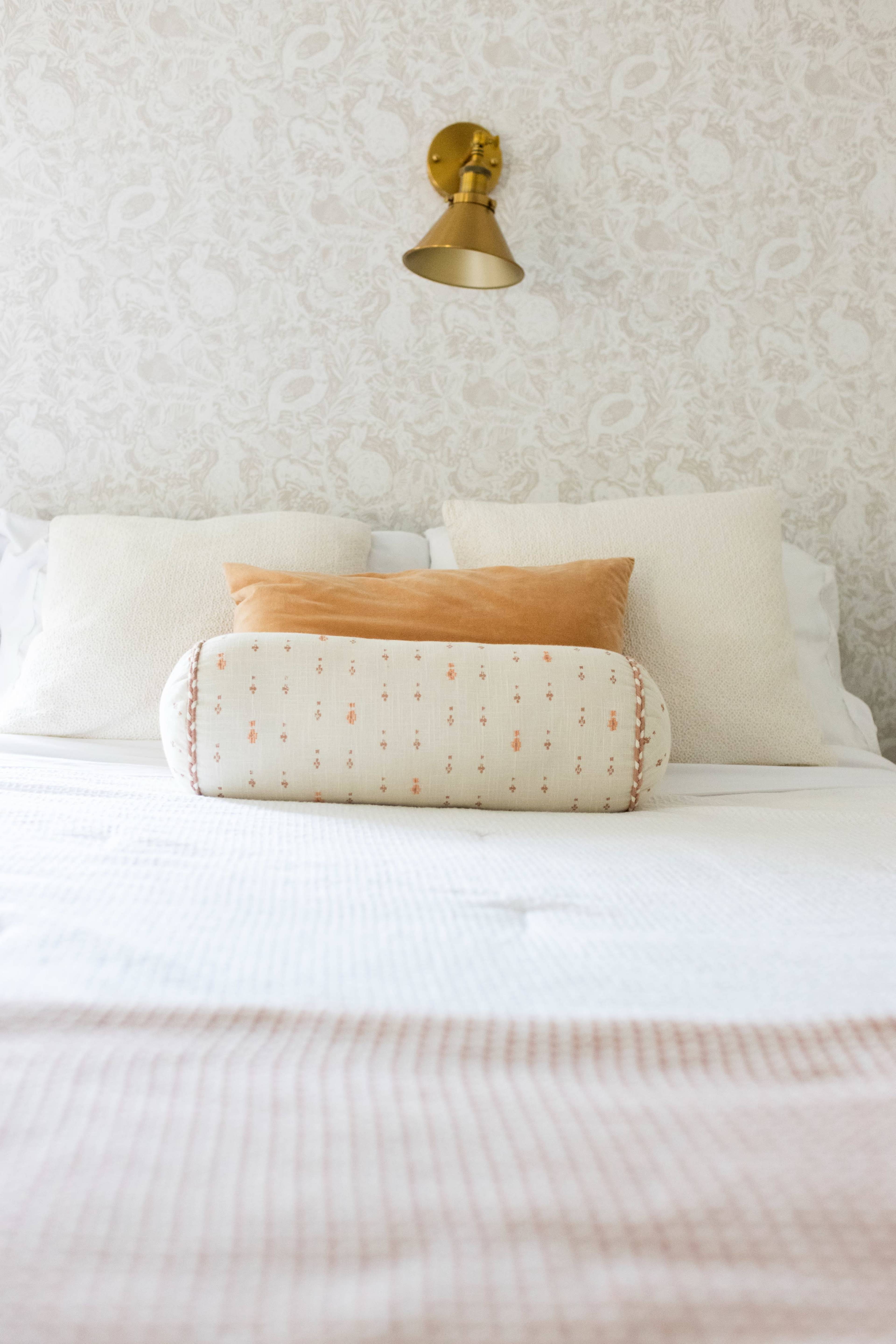 The image shows a neatly made bed with a decorative pillow against a patterned wall and a wall-mounted lamp.