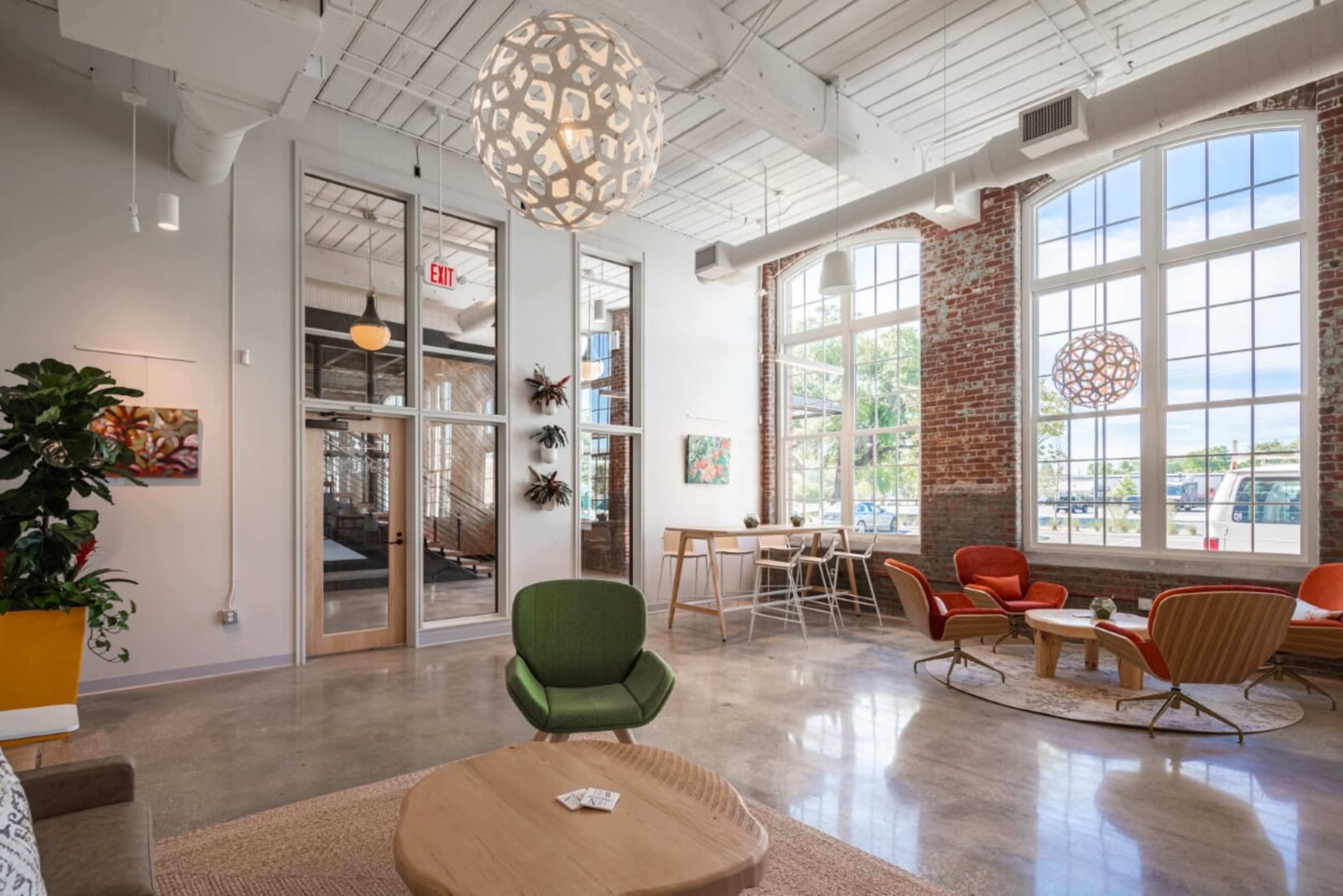 A modern interior space with large windows, exposed brick walls, and a mix of seating arrangements including chairs and a coffee table.