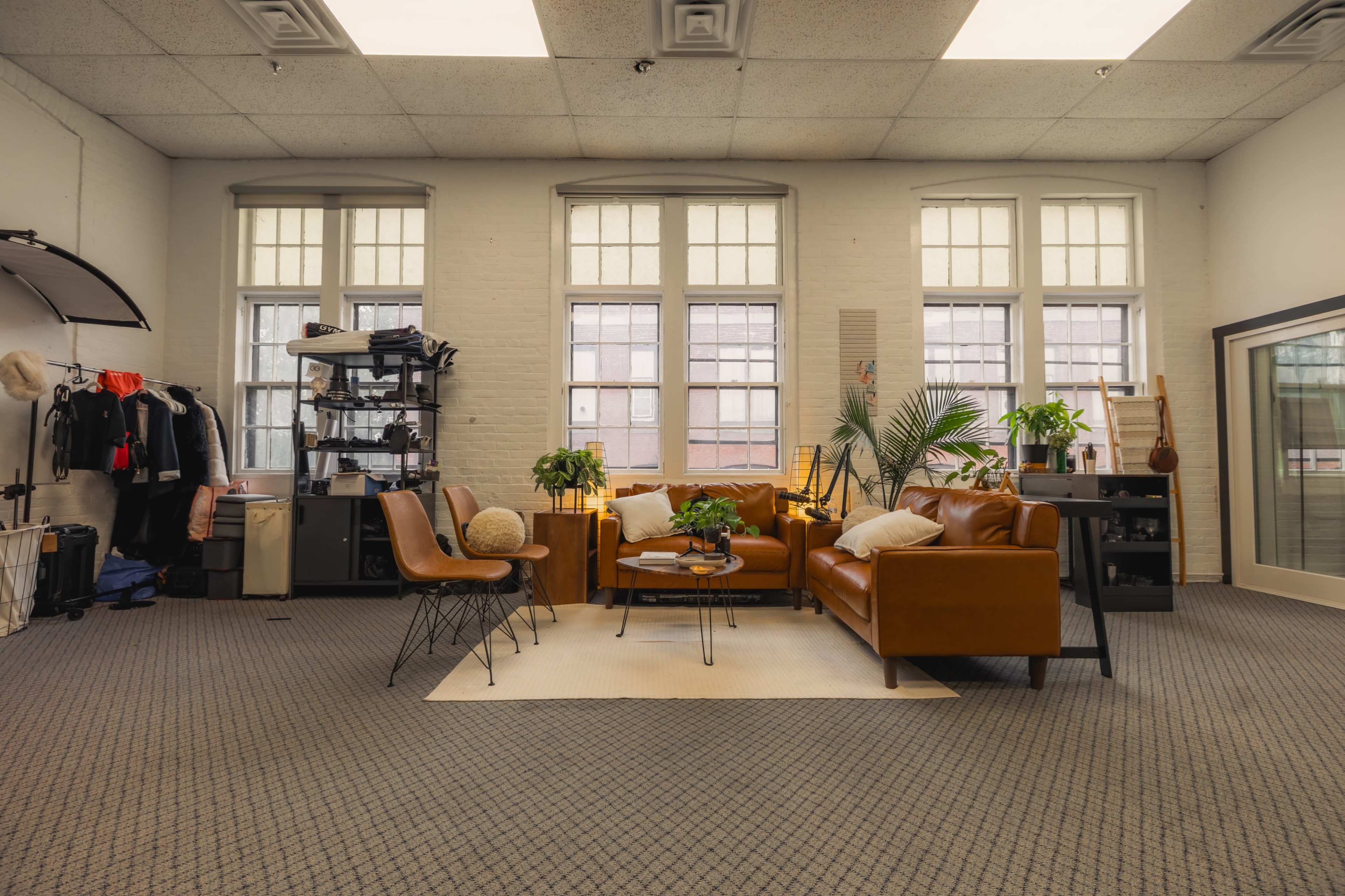 The image shows a well-lit room featuring a seating area with brown leather sofas, a round coffee table, plants, and various items along the walls.