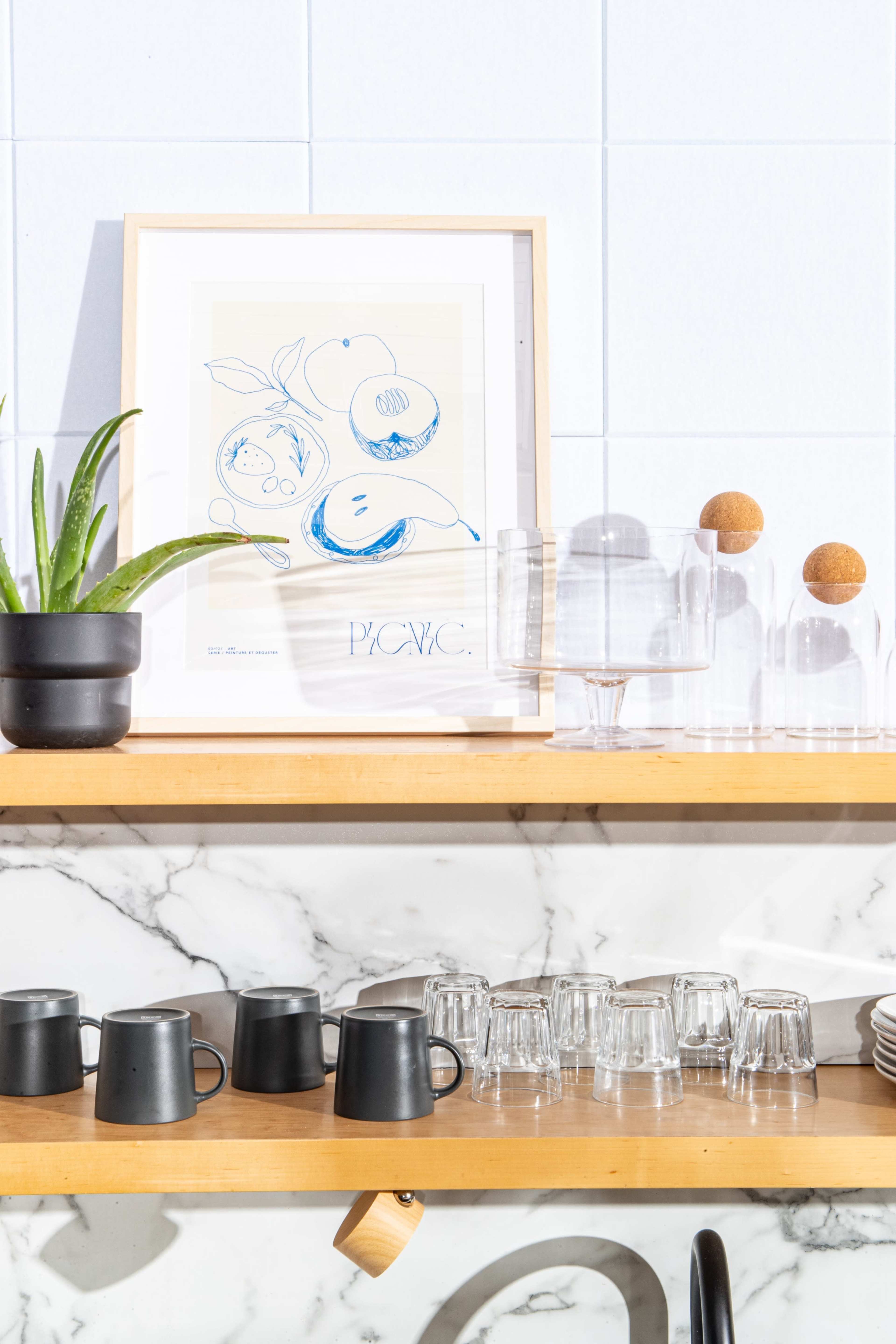 A wooden shelf displays black ceramic mugs and glassware alongside a framed artwork of fruit and a decorative plant.