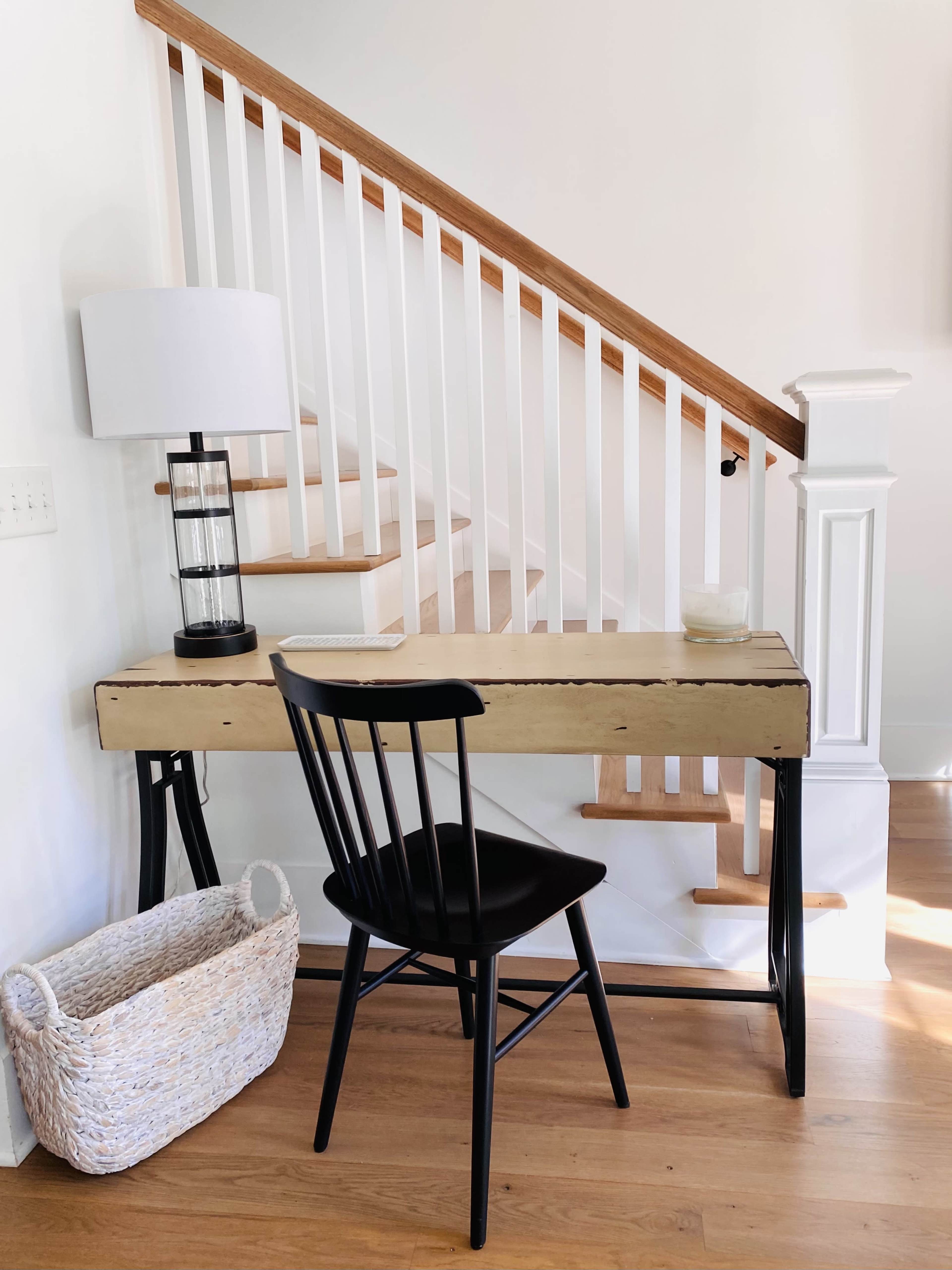 A black chair is positioned at a wooden desk against a white wall near a staircase, with a lamp and a decorative basket nearby.