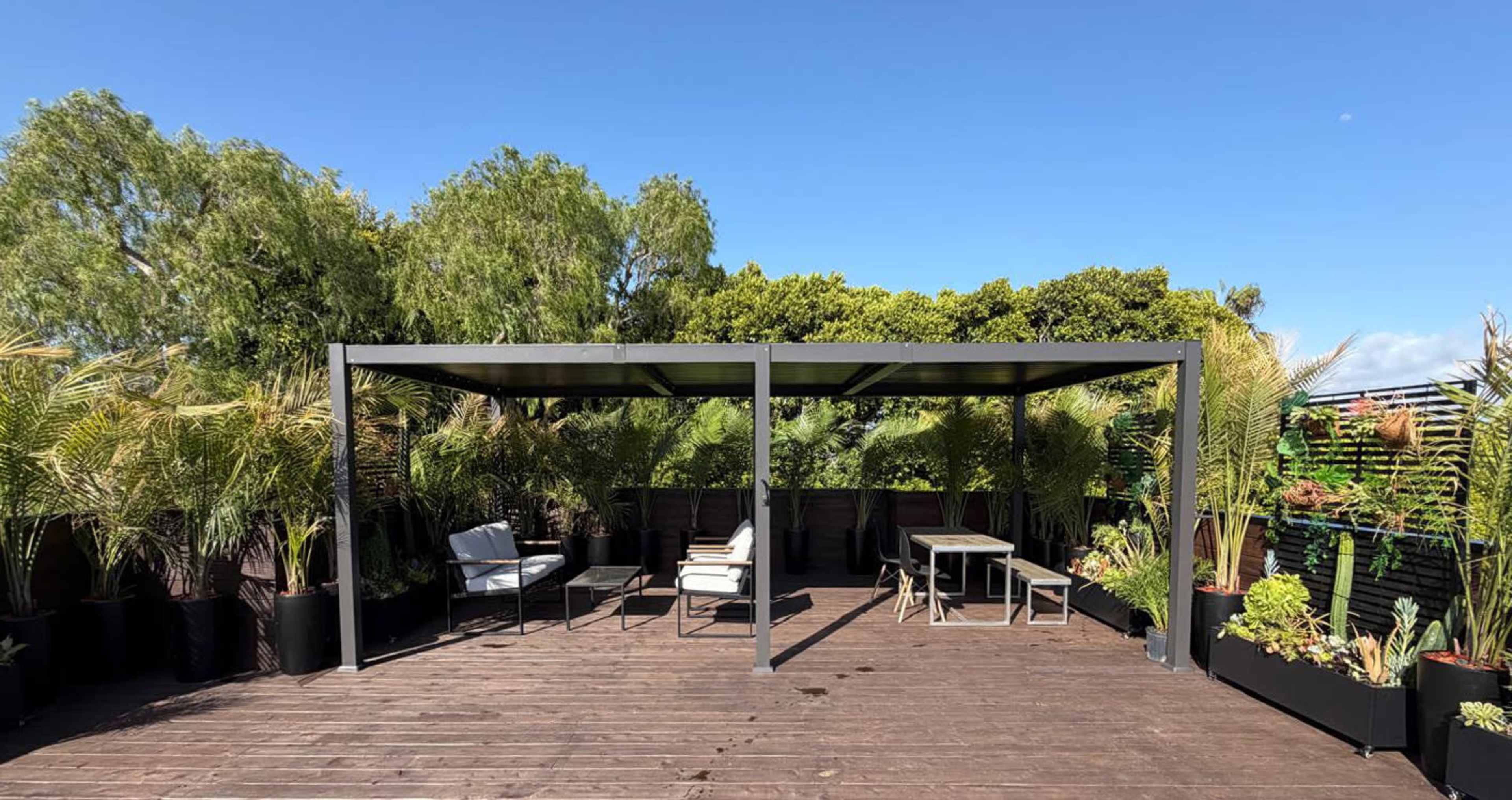 The image shows a rooftop space with a shaded metal structure surrounded by potted plants and seating areas.