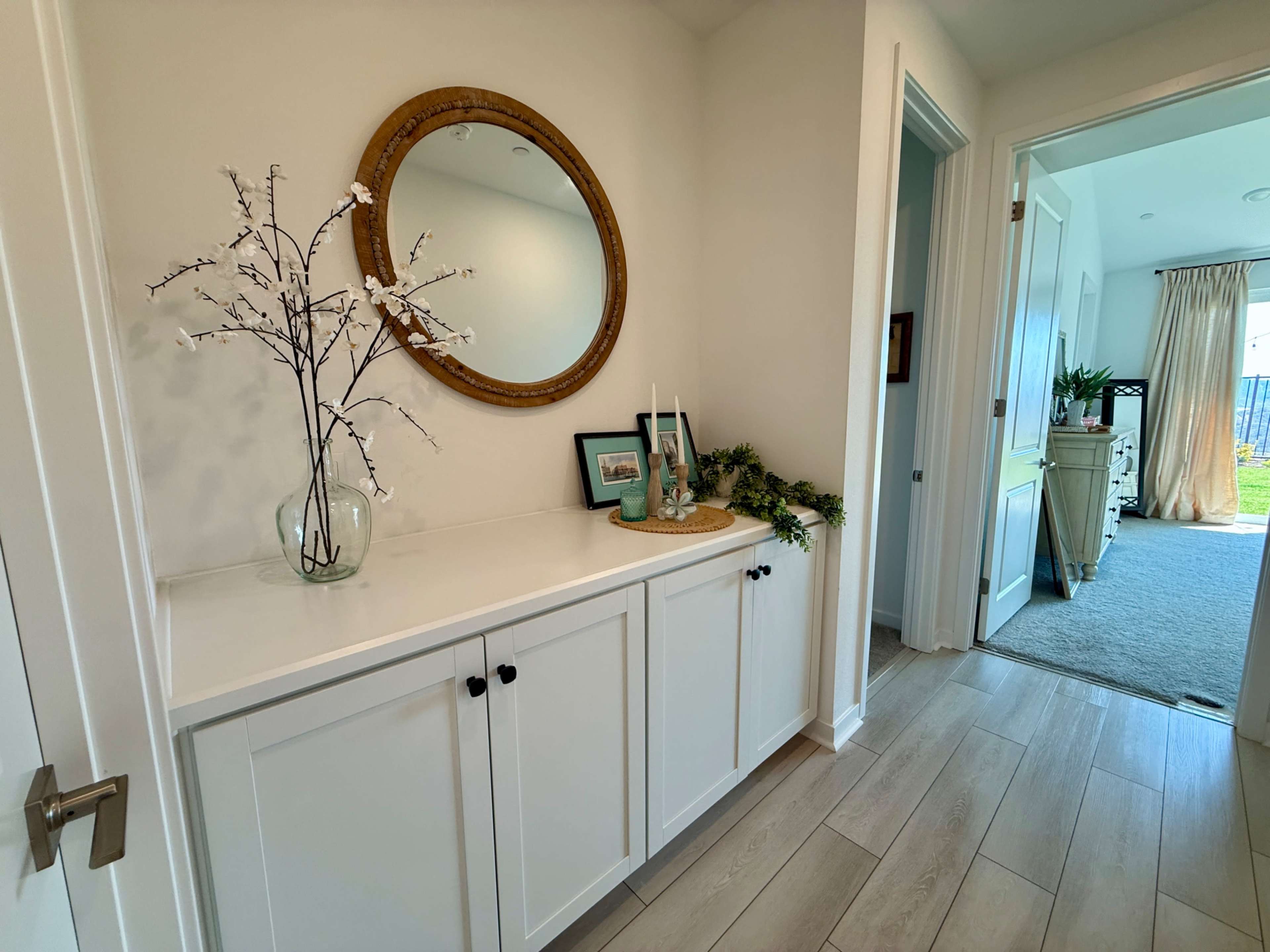 A hallway features a white console table adorned with a decorative mirror, a vase with branches, and framed photos, leading to an open door at the far end.