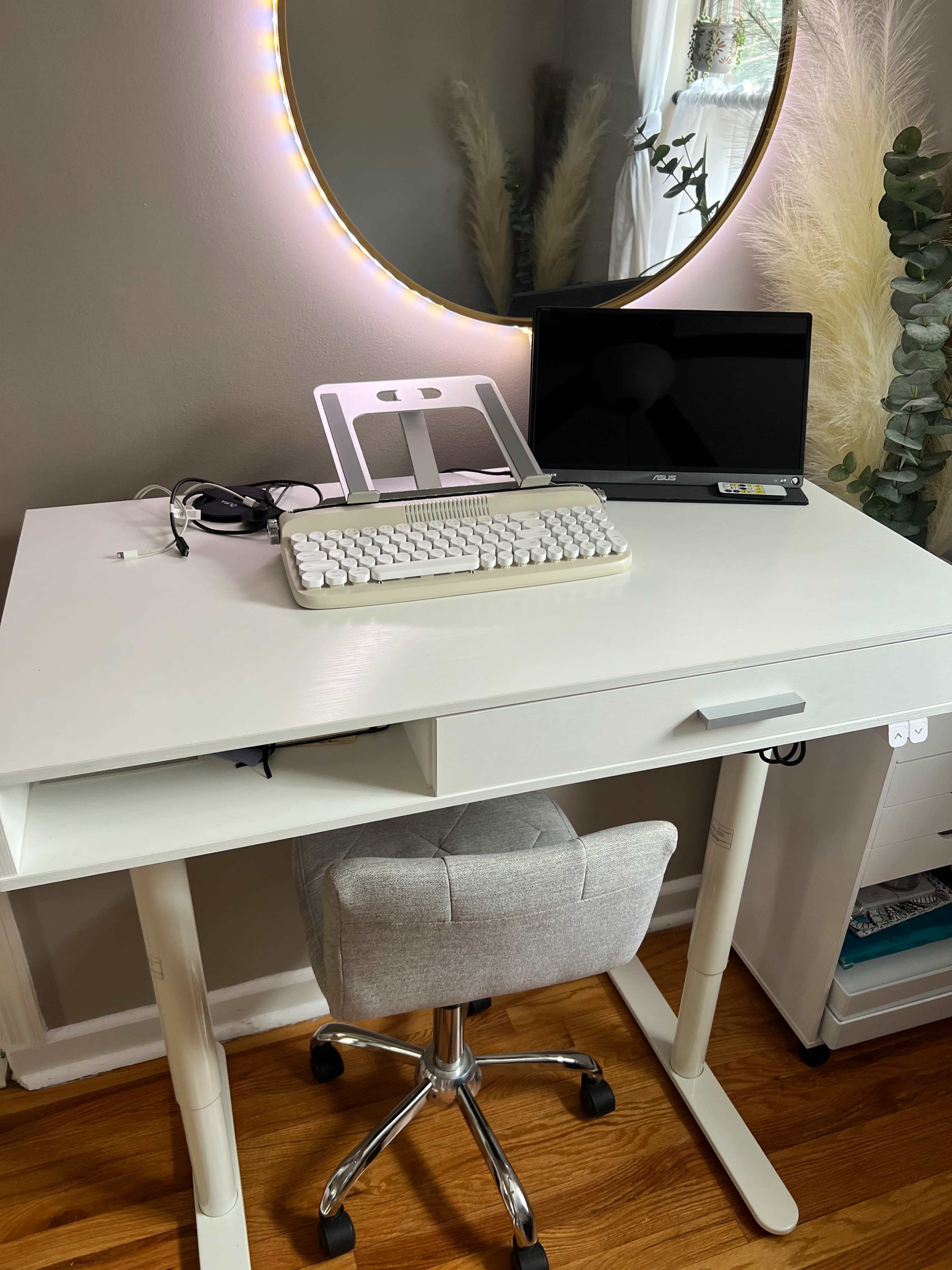 A white desk with a keyboard and a laptop is positioned near a round mirror, surrounded by decorative plants.