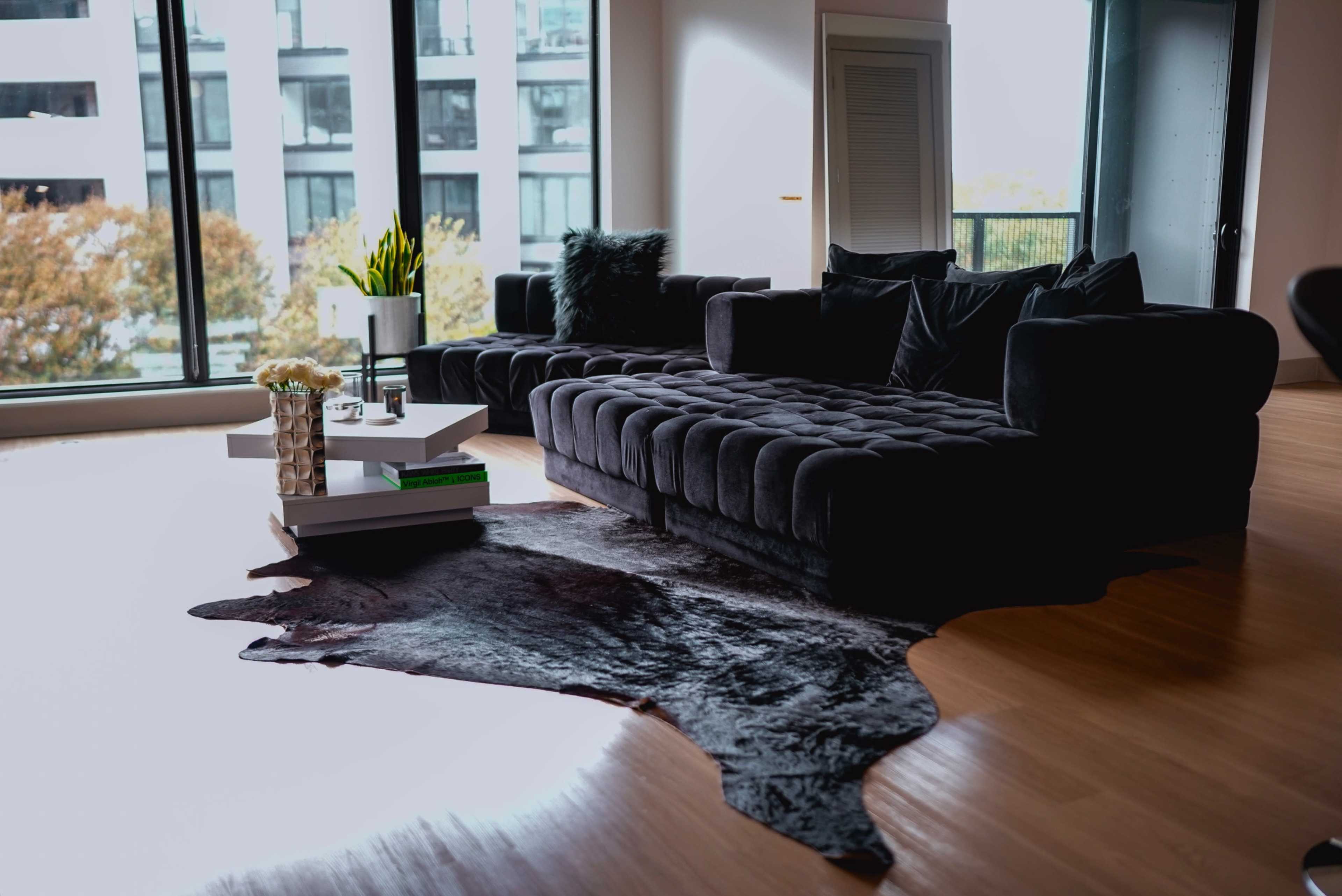 A modern living room featuring a dark sectional sofa, a coffee table with decorative items, and a large area rug on wooden flooring, with large windows providing natural light.