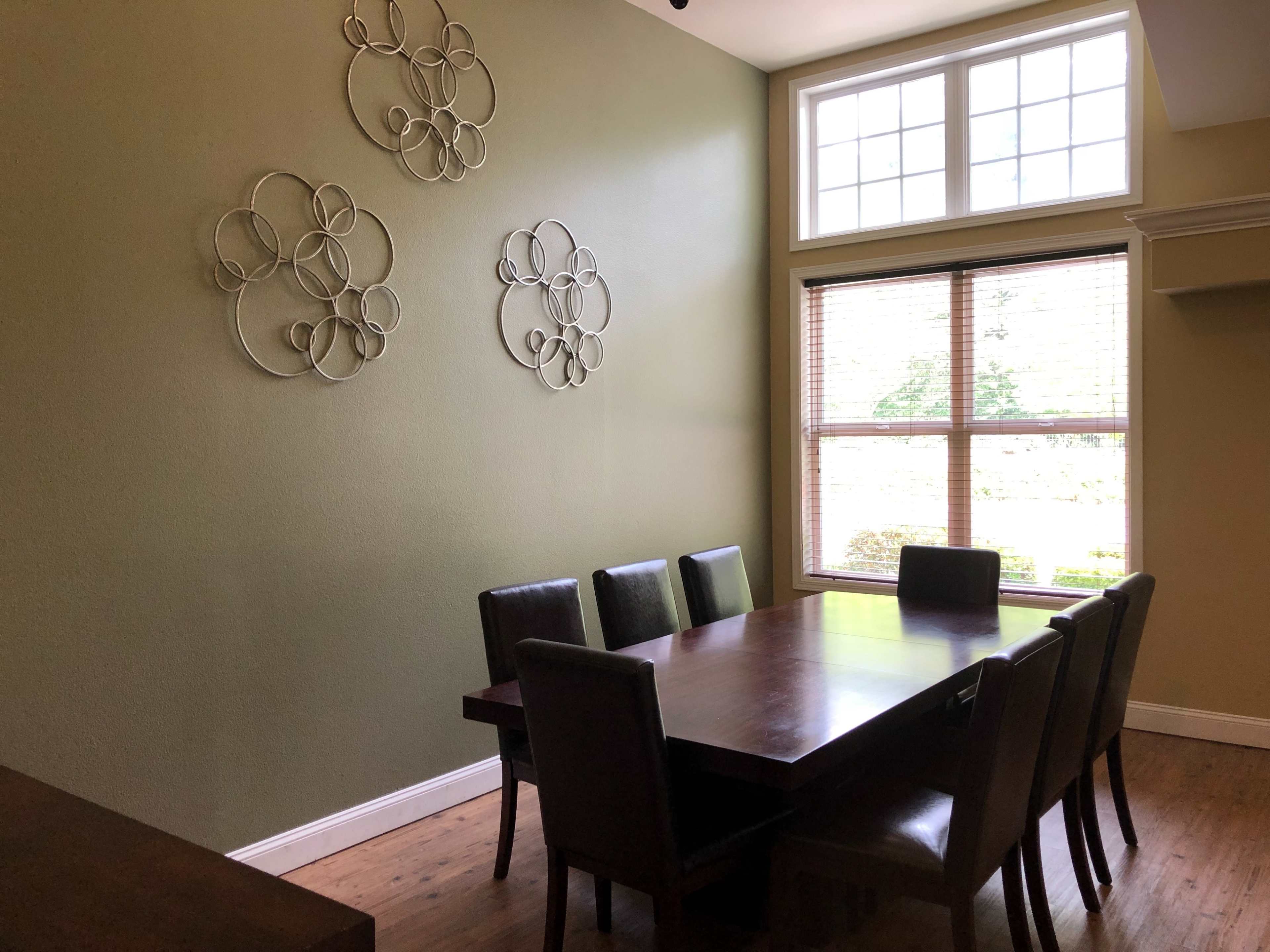 A wooden dining table with six chairs is set against a green wall, featuring decorative metal wall art and a large window allowing natural light to enter.