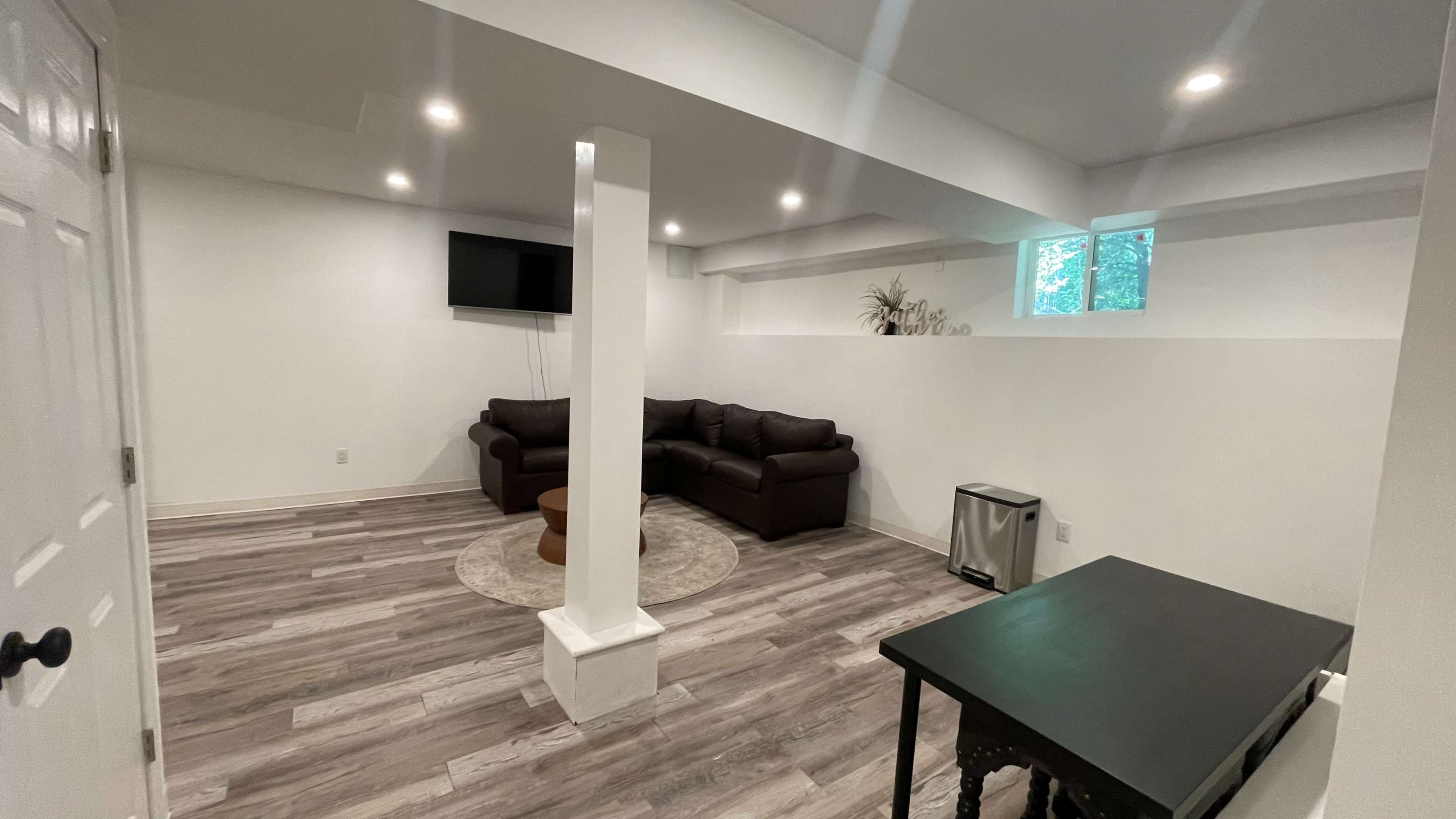 A spacious basement living area featuring a dark couch, a round coffee table, a television mounted on the wall, and a small trash bin in a corner.