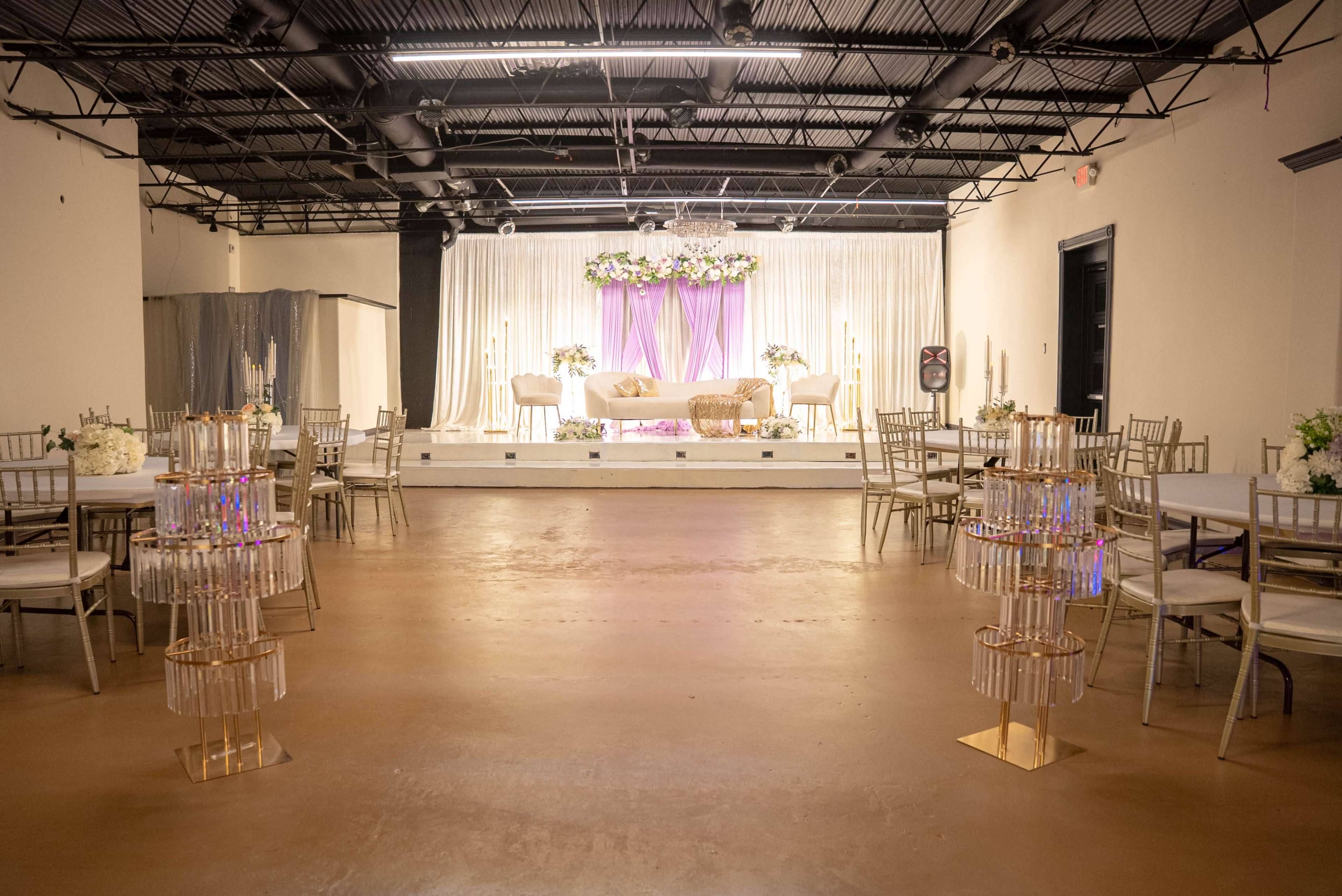 The image shows an empty event hall with gold and white decor, featuring tables and chairs arranged around a stage adorned with floral arrangements and a purple backdrop.
