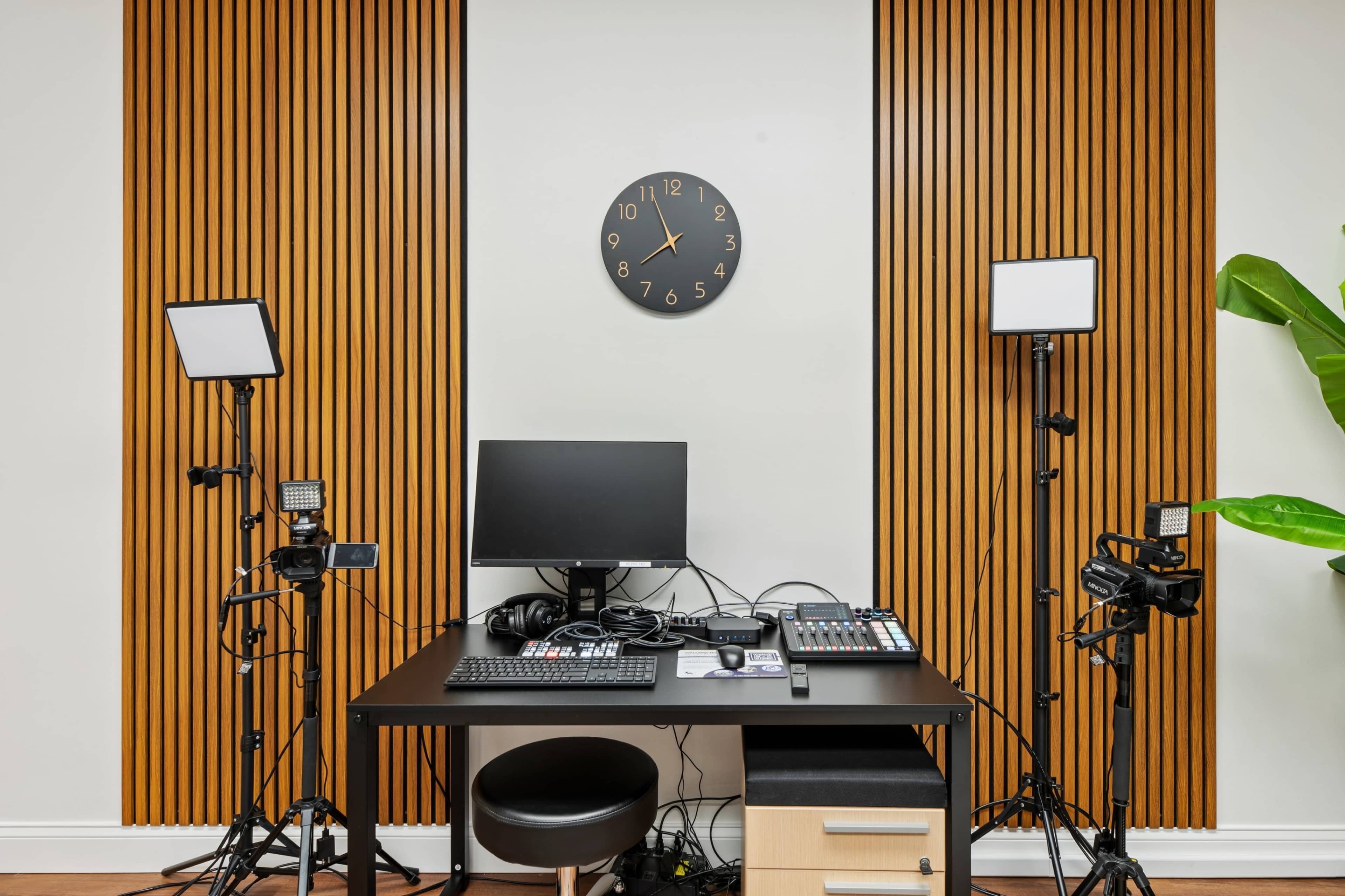 A minimalistic studio setup features a desk with a computer, video control equipment, and lighting on either side, against a wall with vertical wooden slats and a clock above.