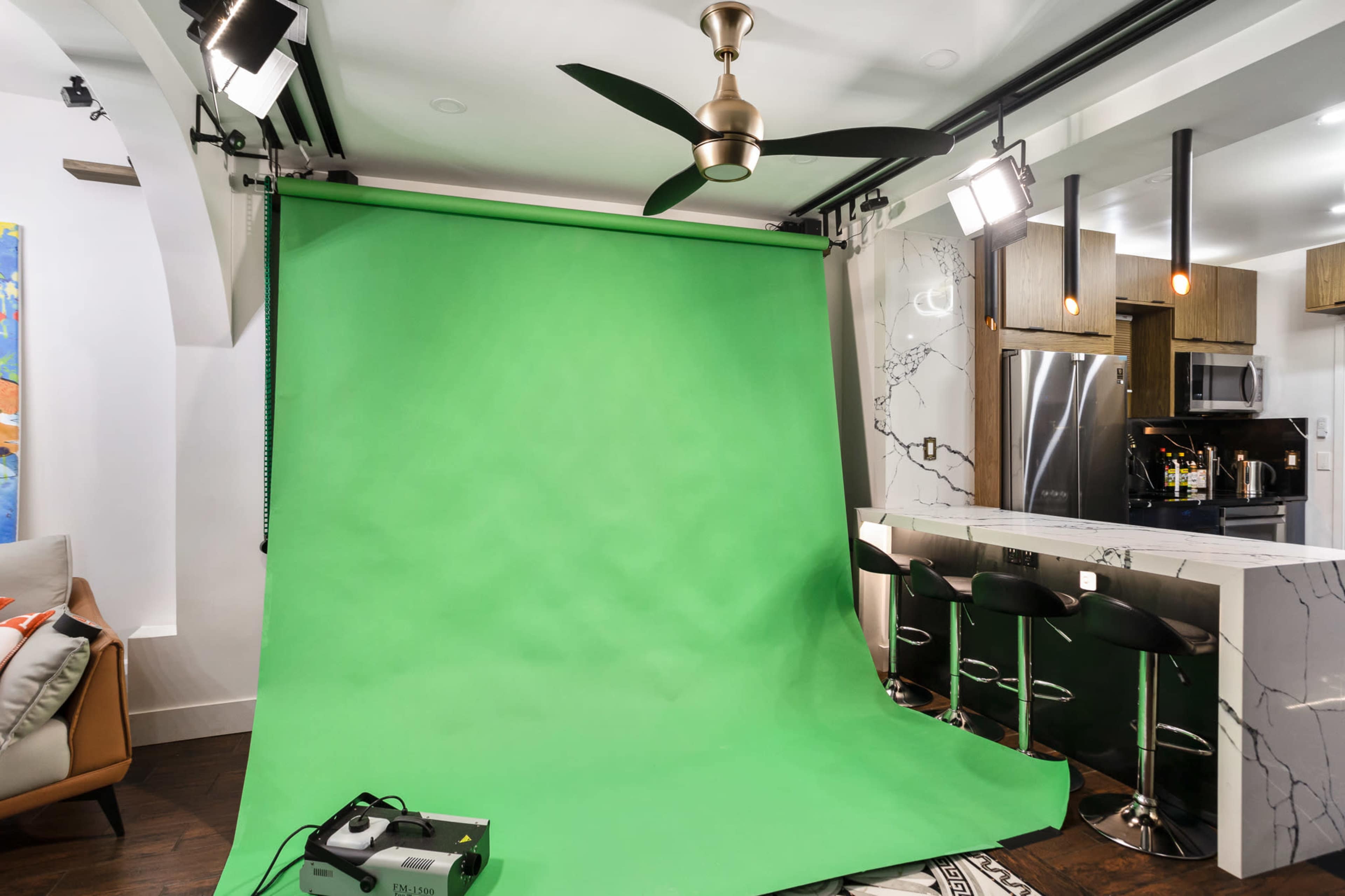 A studio setup featuring a green backdrop, a ceiling fan, and kitchen appliances visible in the background.