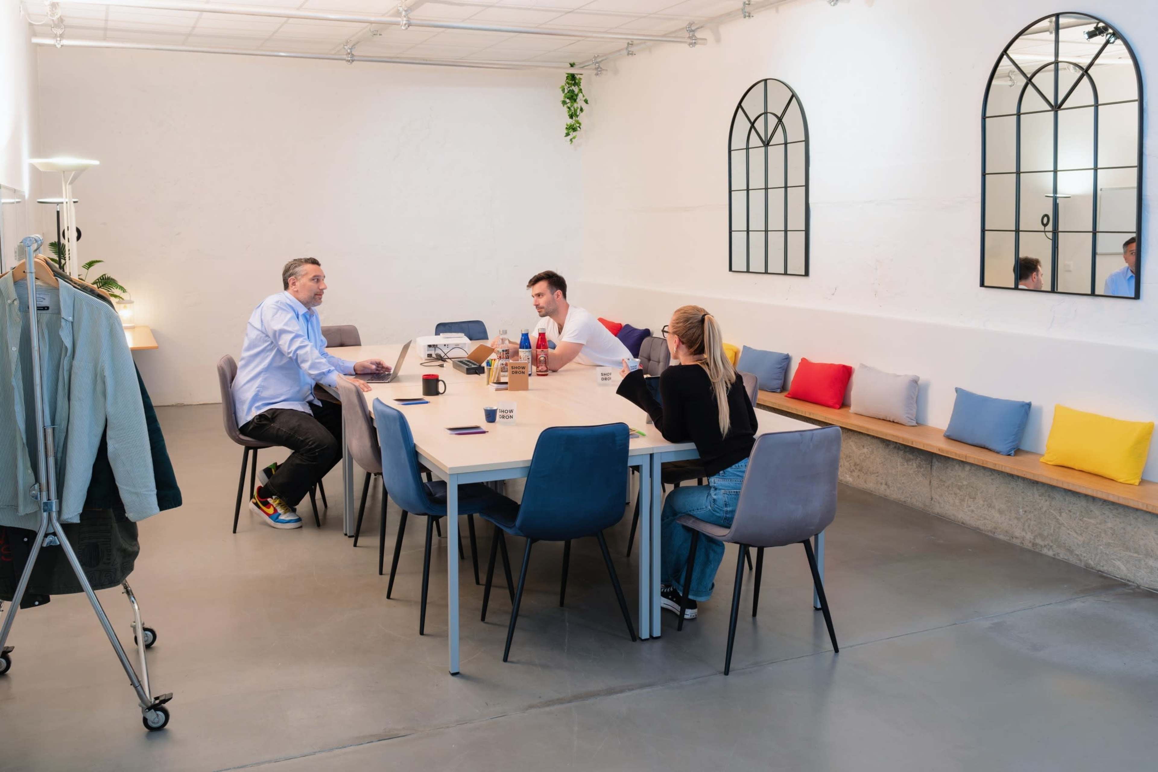 A group of three people is seated around a long table in a bright, modern meeting room with white walls and colorful cushions along a bench.