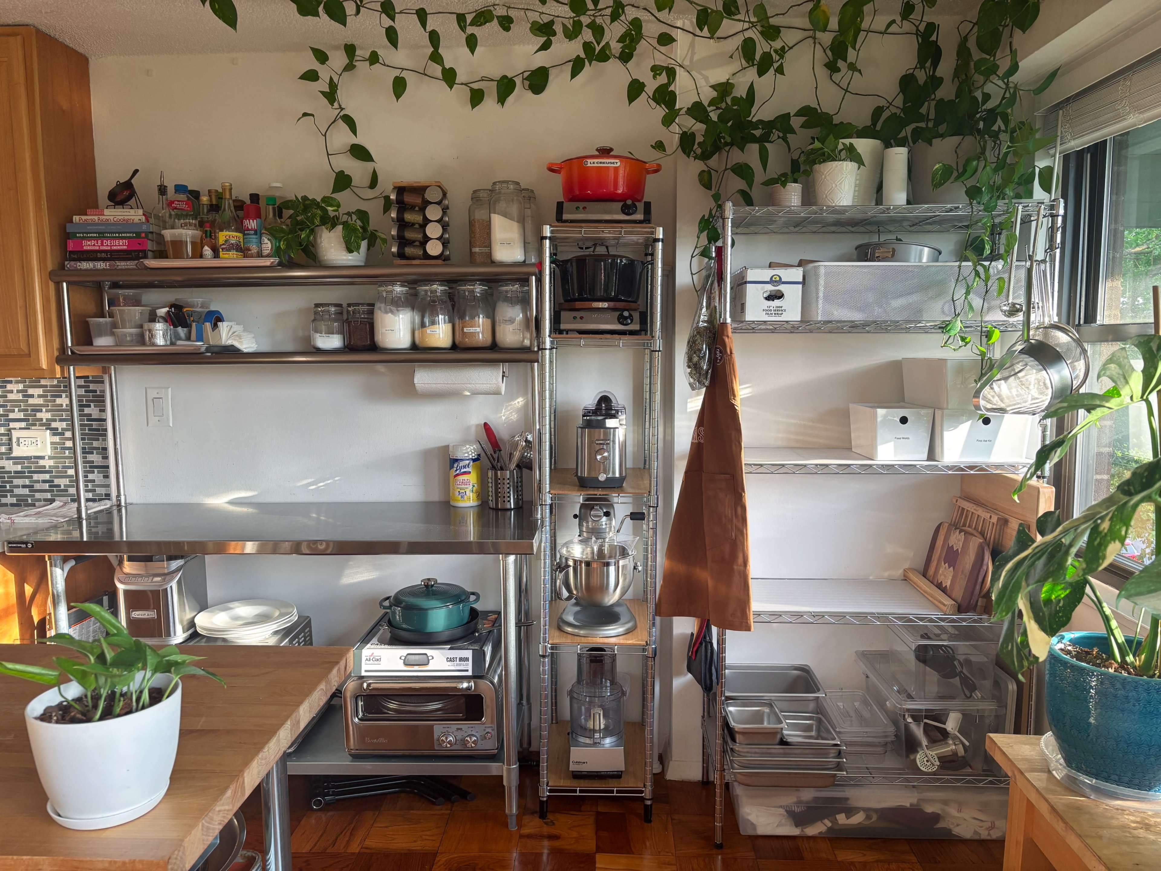 The image shows a tidy kitchen corner with metal shelving units filled with jars, kitchen appliances, and plants, alongside a wooden countertop.