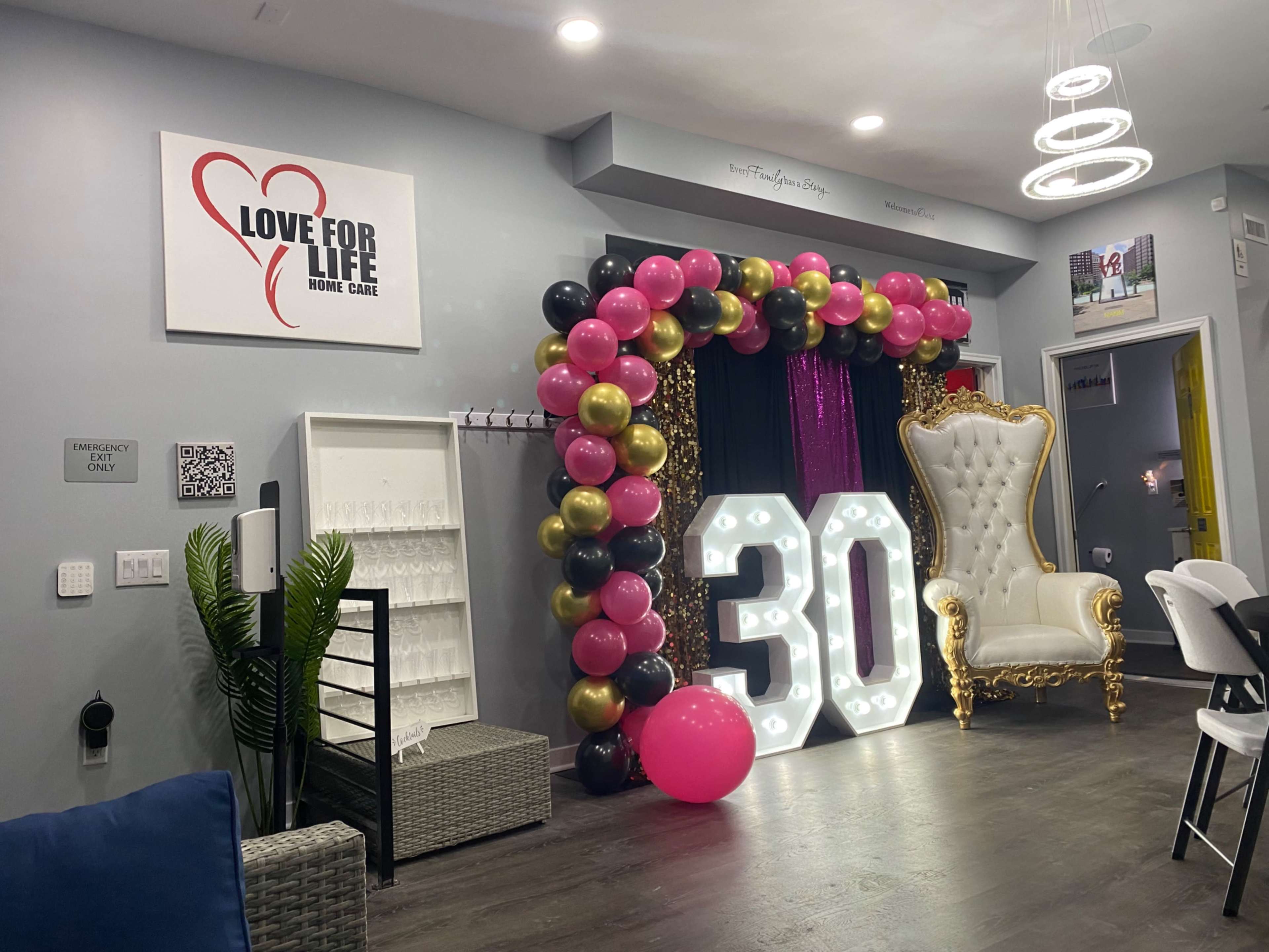 The image shows a decorated room featuring a large gold-framed chair, a backdrop of pink and black balloons, and illuminated numbers reading "30."
