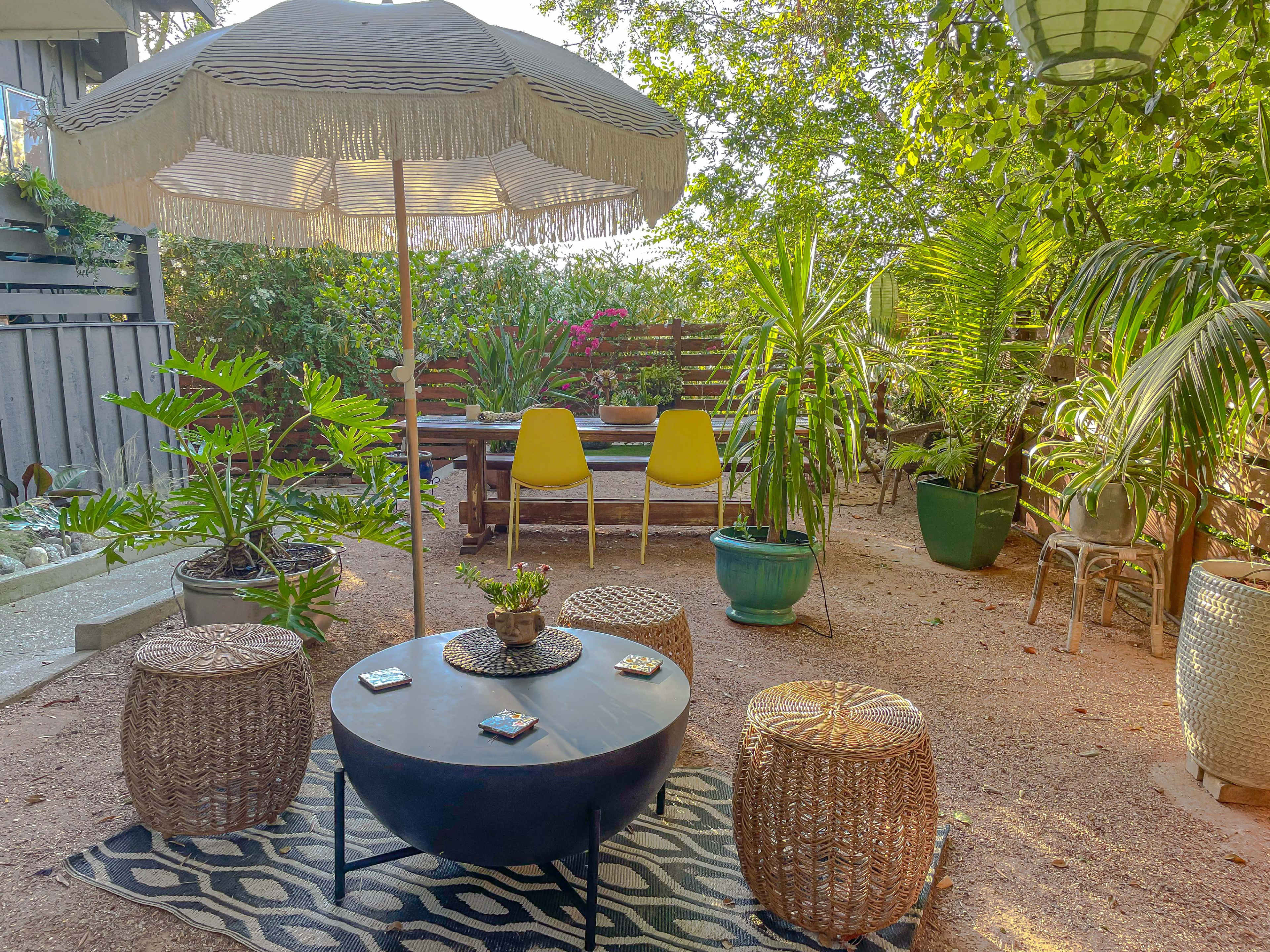 A shaded seating area with a large umbrella features a round table surrounded by woven stools and potted plants in a backyard garden.