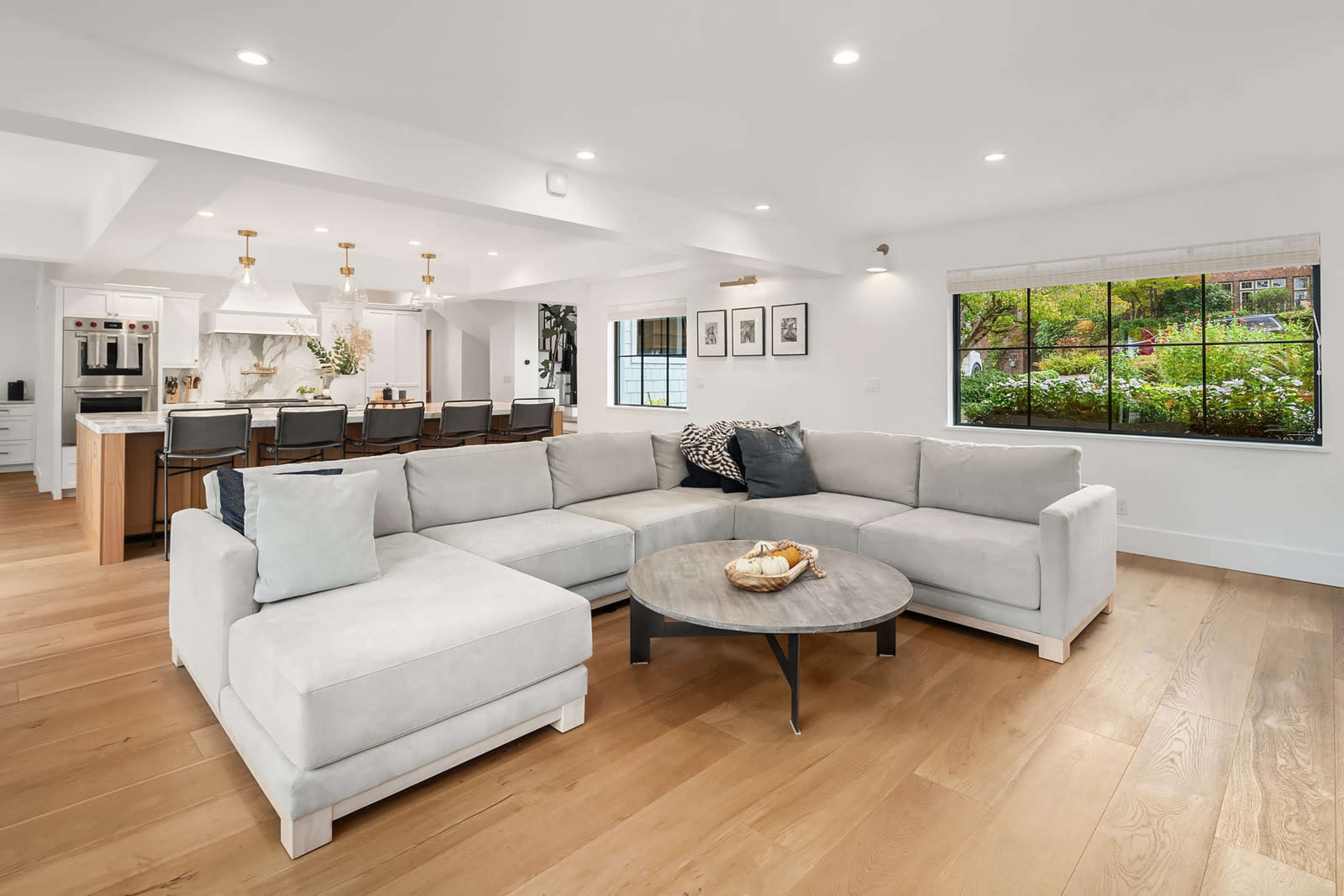 A spacious living room features a large sectional sofa and a round coffee table, with a view of greenery through a window.