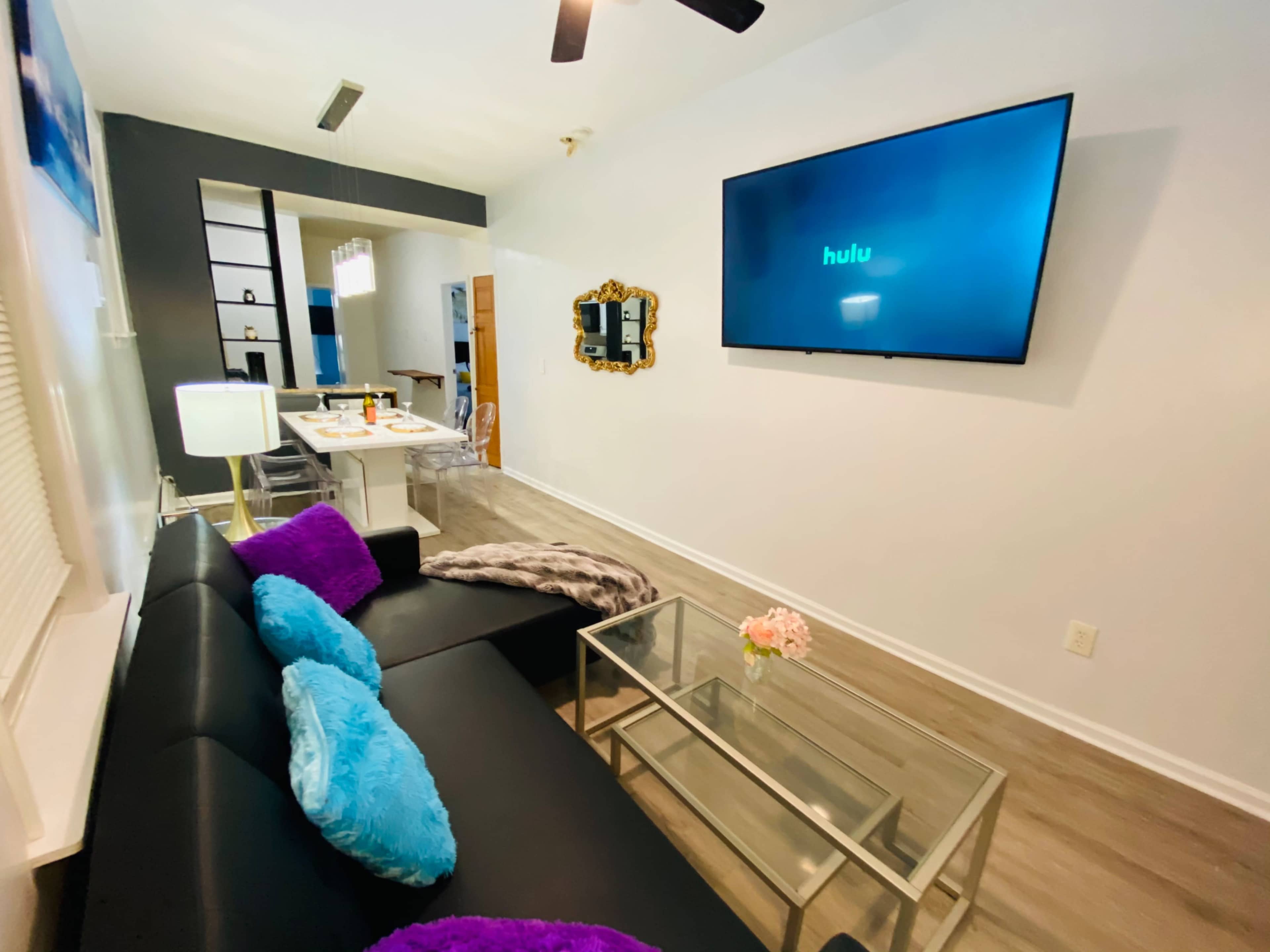 The image shows a modern living room with a black couch, a glass coffee table, and a wall-mounted TV displaying the Hulu logo.
