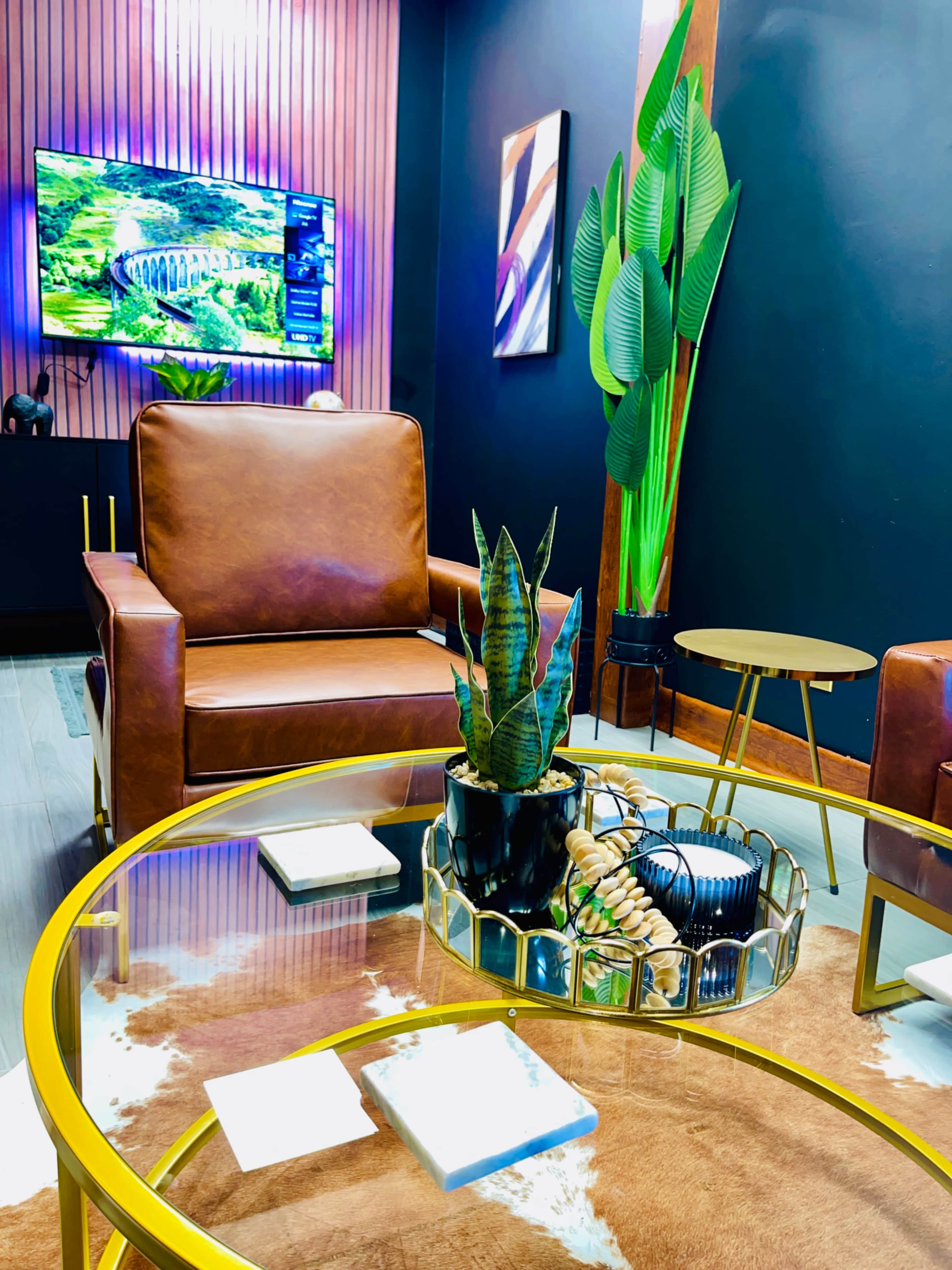 A modern living space featuring brown leather chairs, a glass coffee table with decorative items, and a large television mounted on the wall.