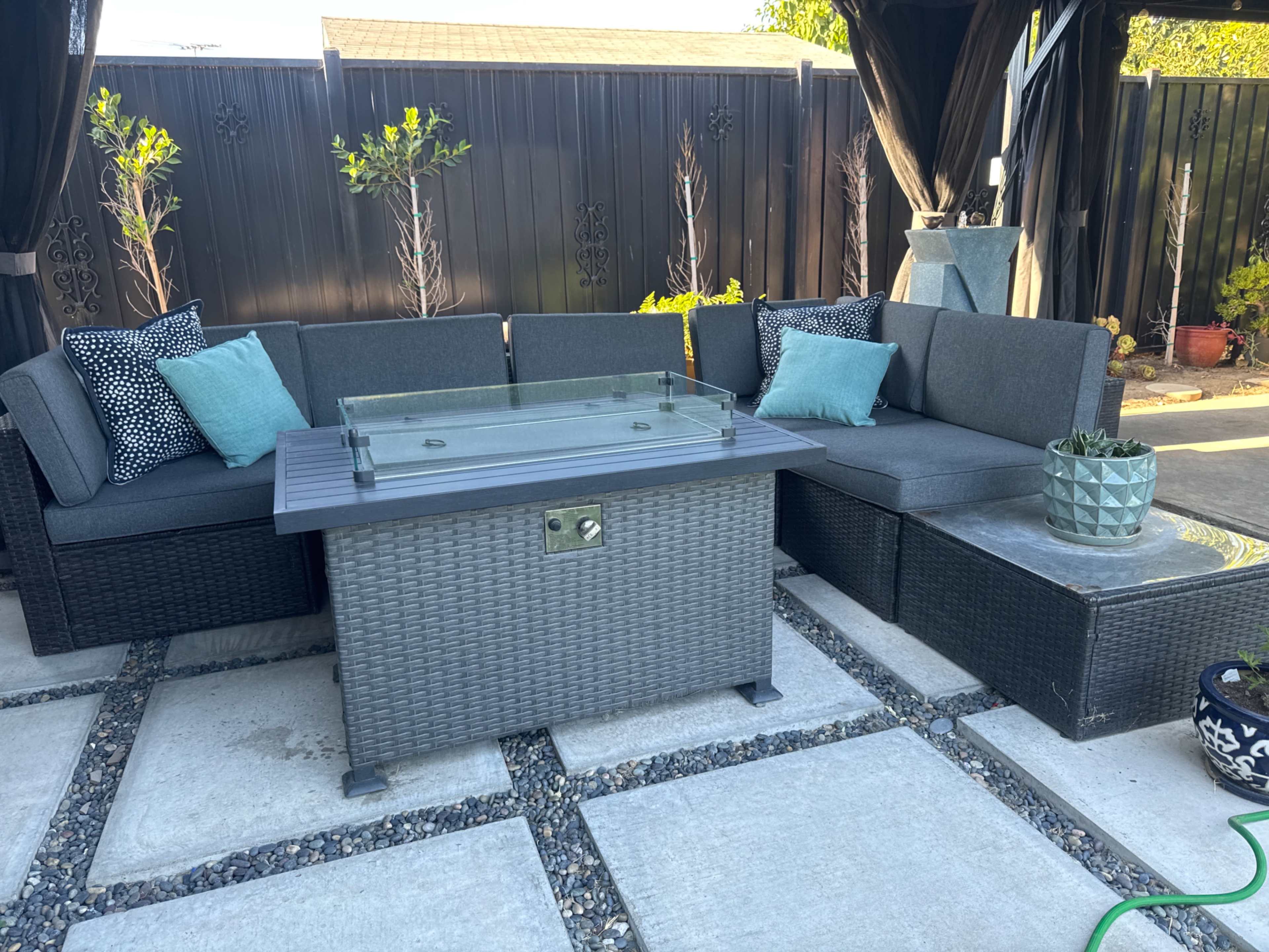 The image shows a backyard seating area with a sectional sofa, a fire pit table in the center, and decorative plants nearby.