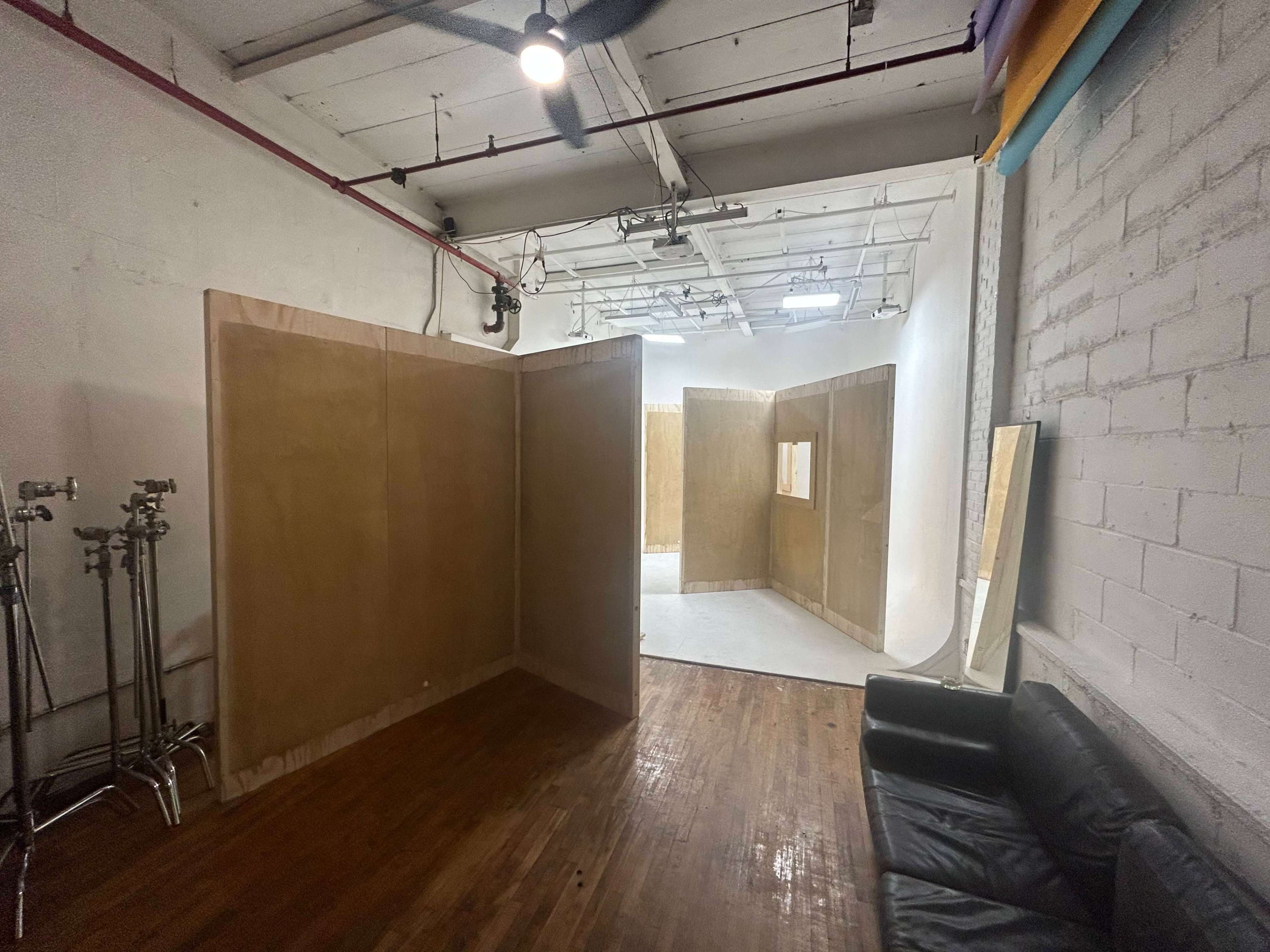 The image shows an industrial-style studio space with wooden partitions, a simple backdrop area, and a black couch against the wall.