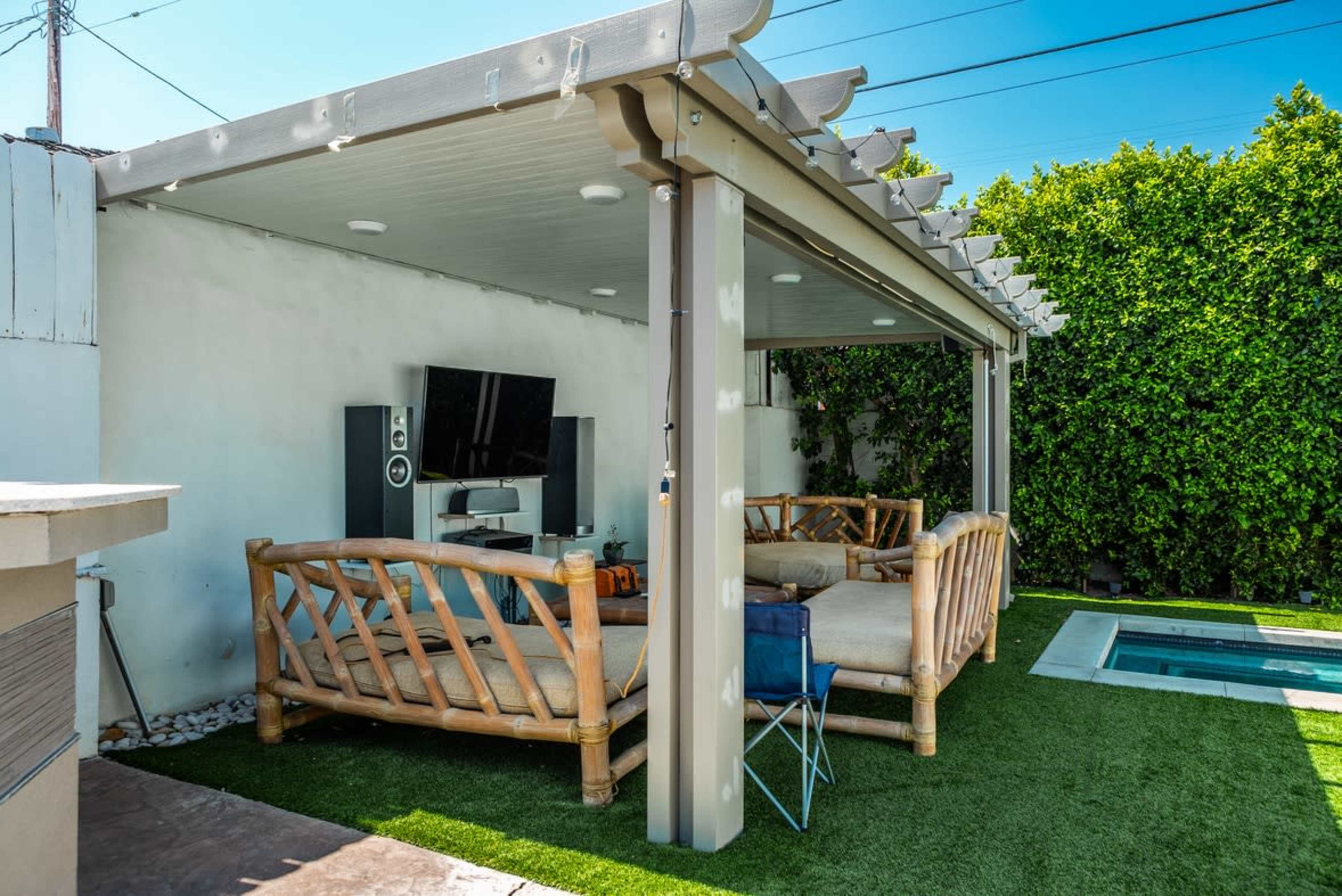 A backyard patio area with bamboo-style furniture, a television mounted on the wall, and a small pool nearby, all surrounded by greenery.