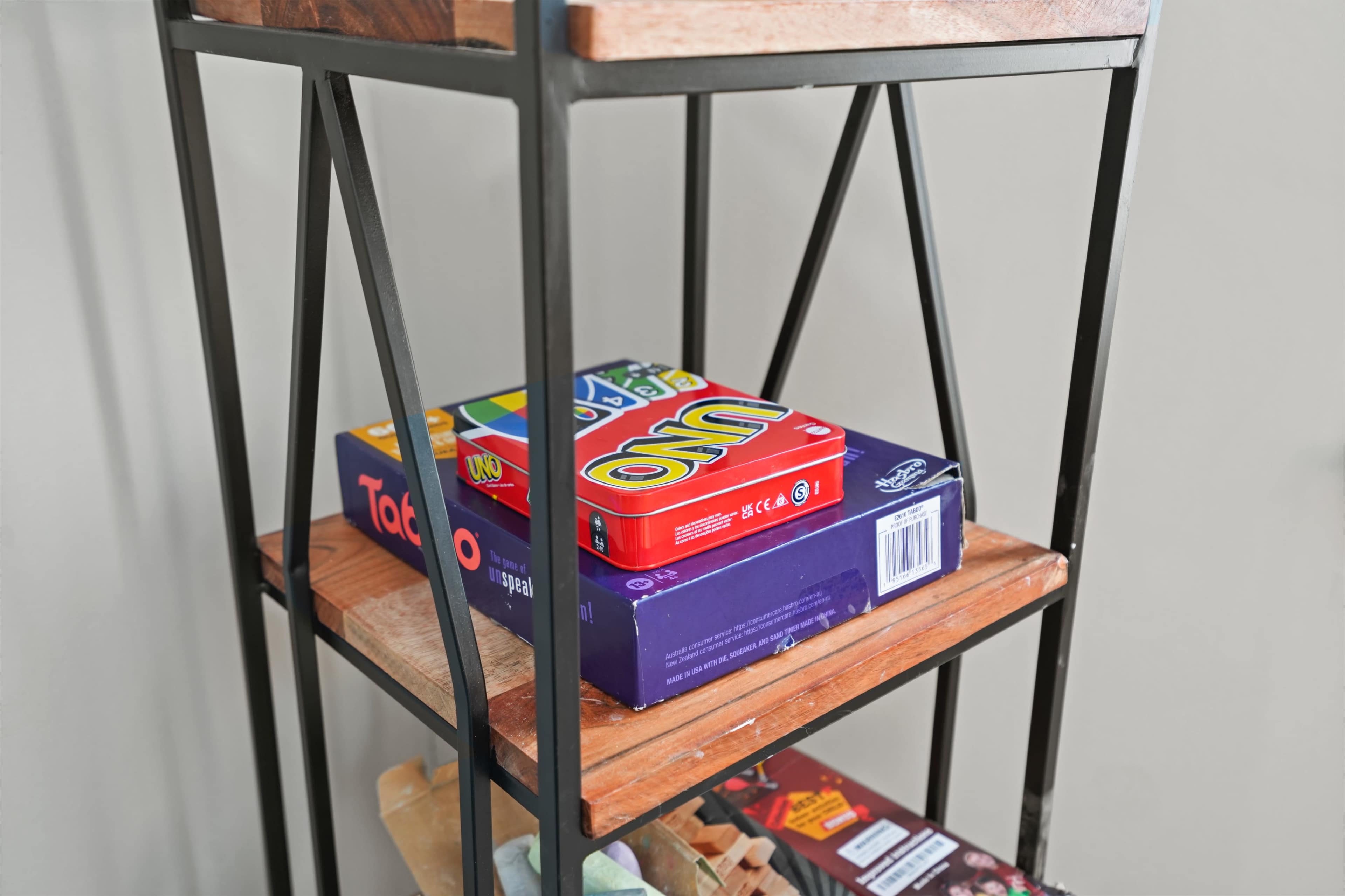 A metal-framed shelf with wooden shelves holds a stack of board games, including Uno and Taboo.