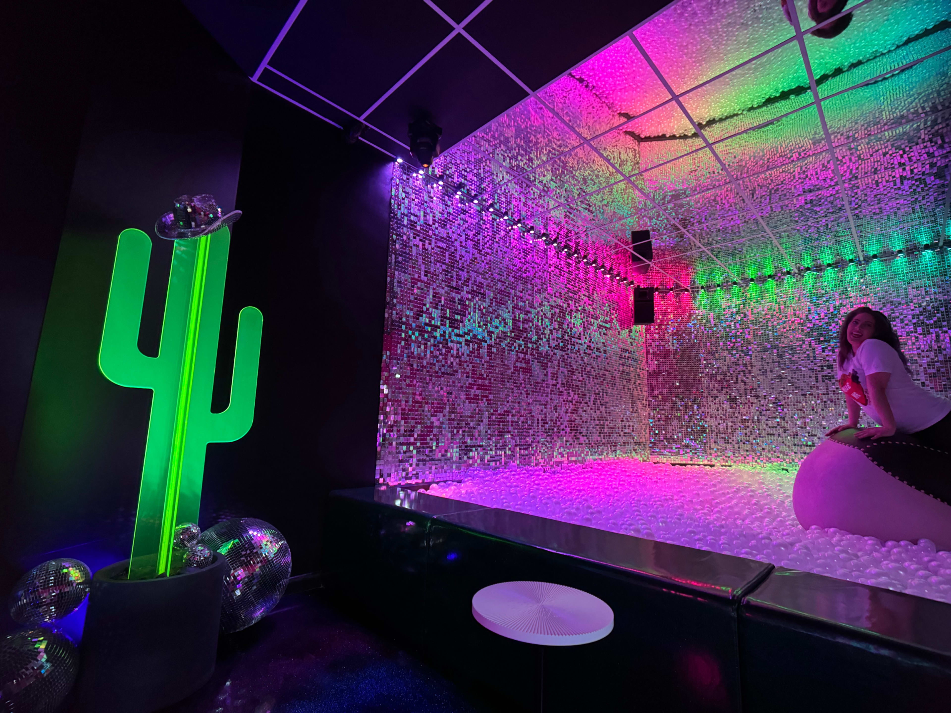 The scene features a brightly colored room with a mirrored wall, a large cactus sculpture, and a woman sitting on a large inflatable object surrounded by a ball pit.