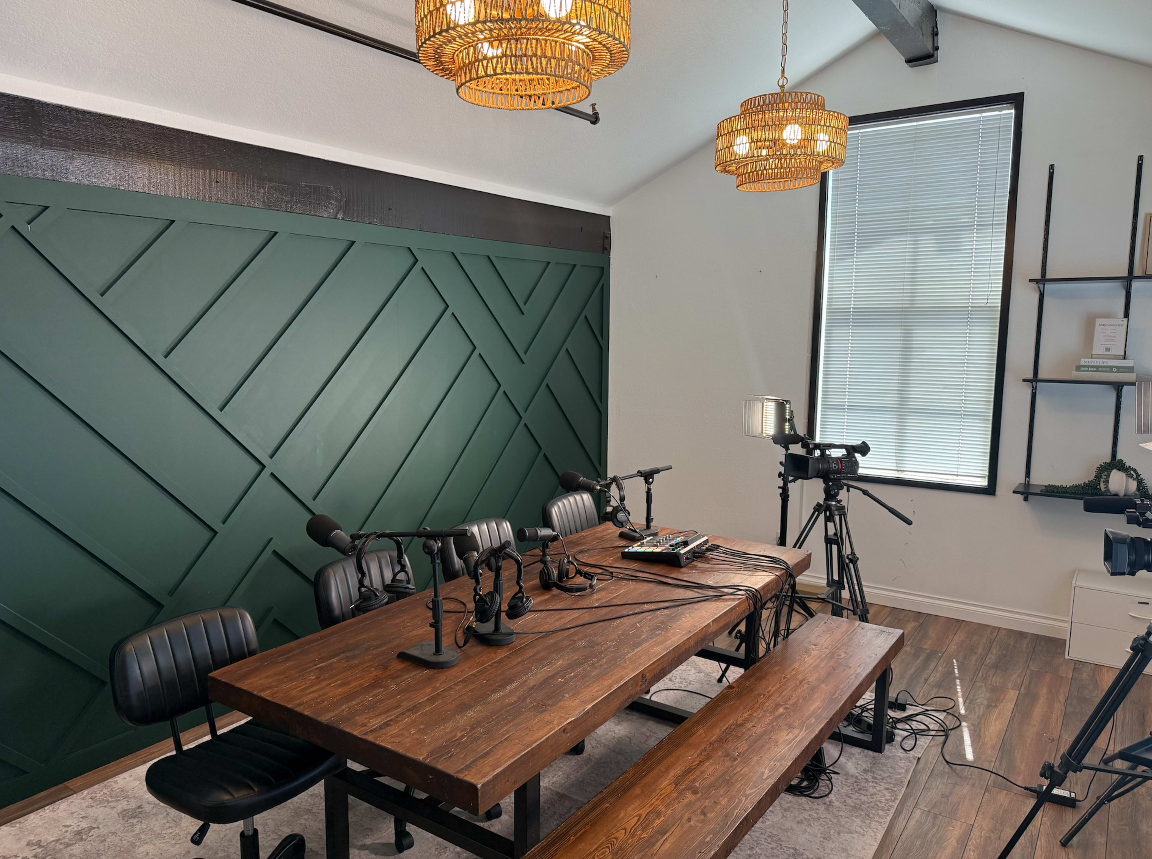 The image shows a podcasting studio with a wooden table, multiple microphones, cameras, and a green paneled wall.