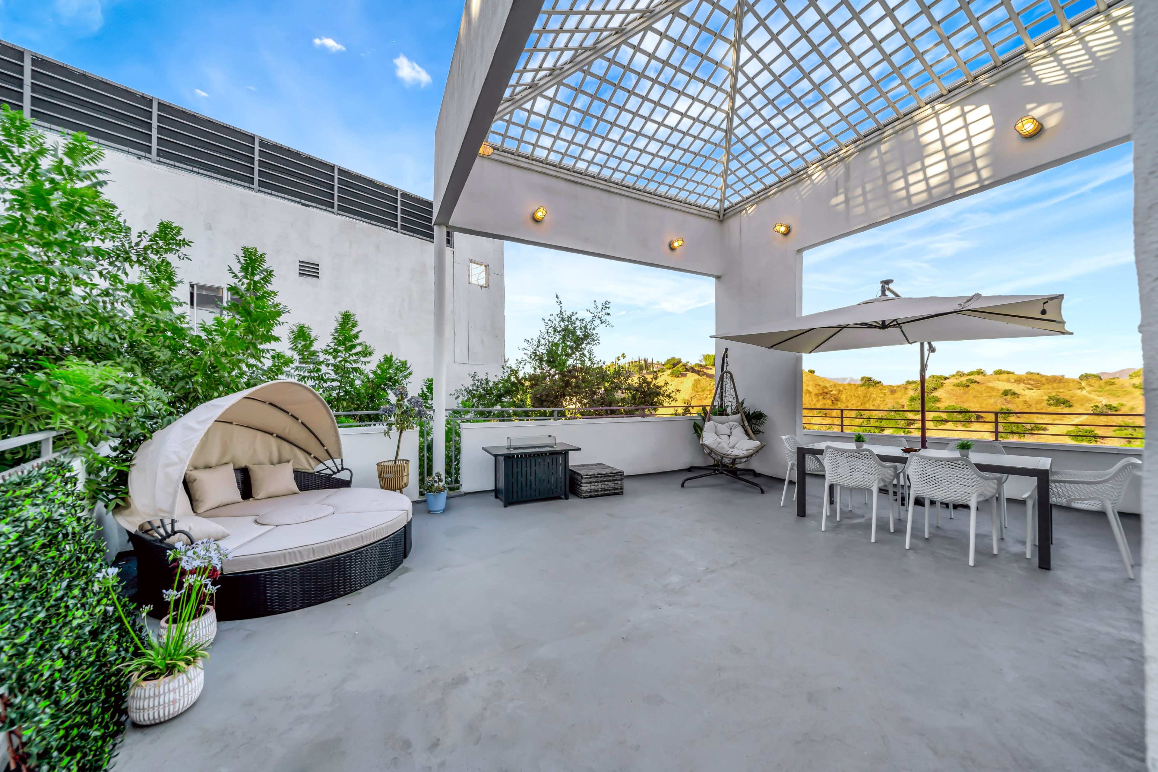 A spacious rooftop terrace featuring a circular lounging area, a dining table under an umbrella, and greenery along the periphery.