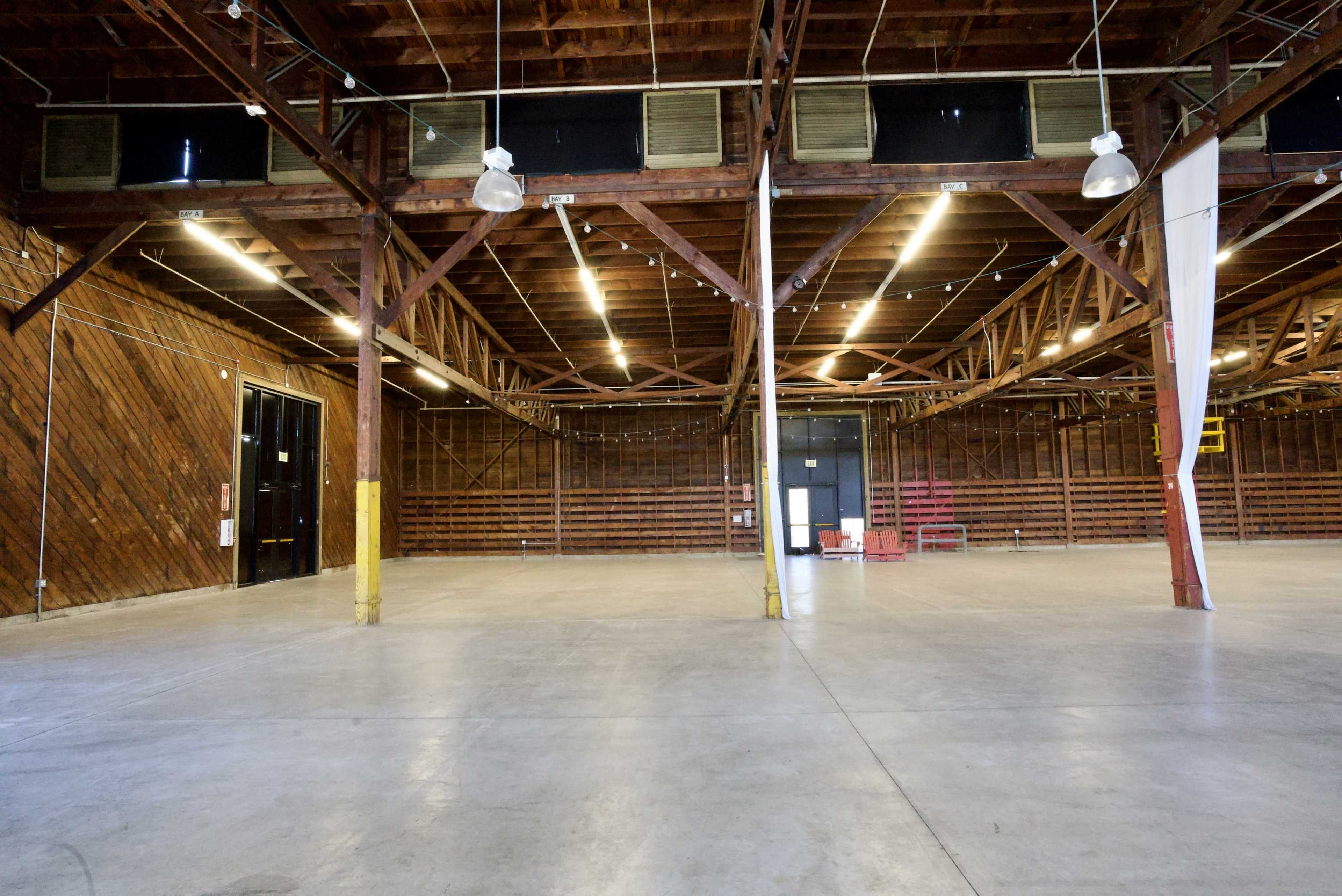 The image shows a large, empty warehouse space with wooden beams and a concrete floor.